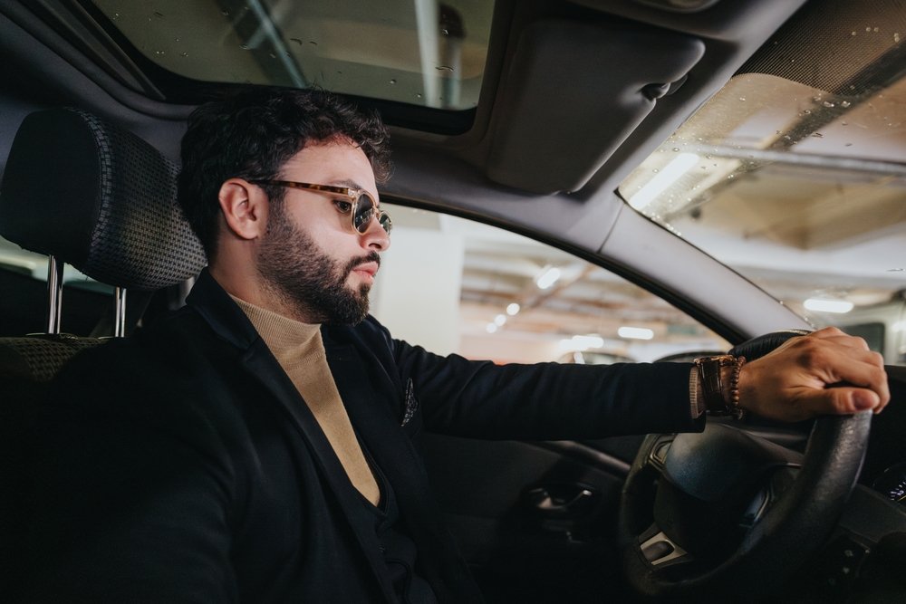 Man with sunglasses and beard driving a car in an indoor parking garage.