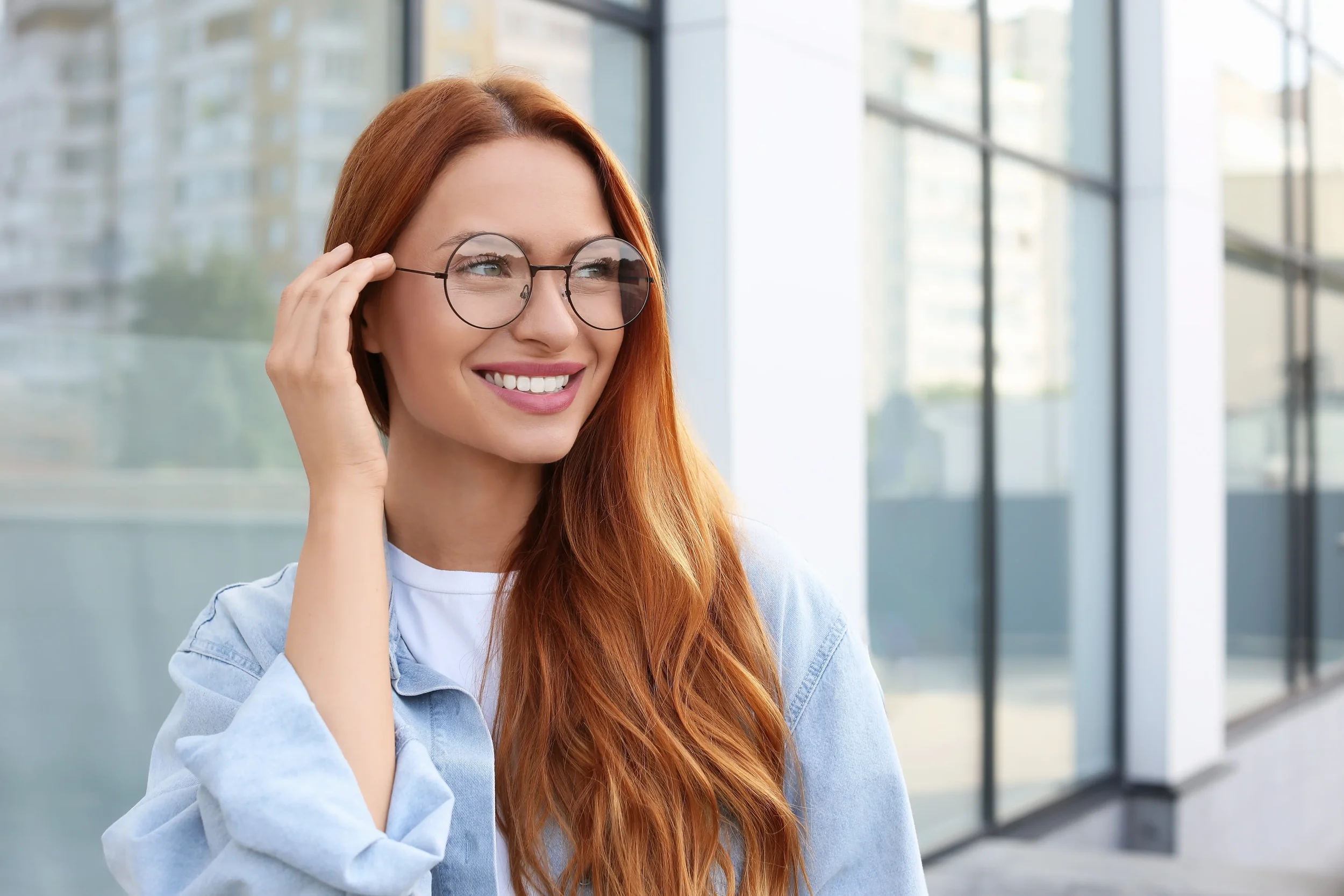 A young woman with long red hair and glasses smiling and adjusting her glasses, standing in front of large glass windows in a modern building.