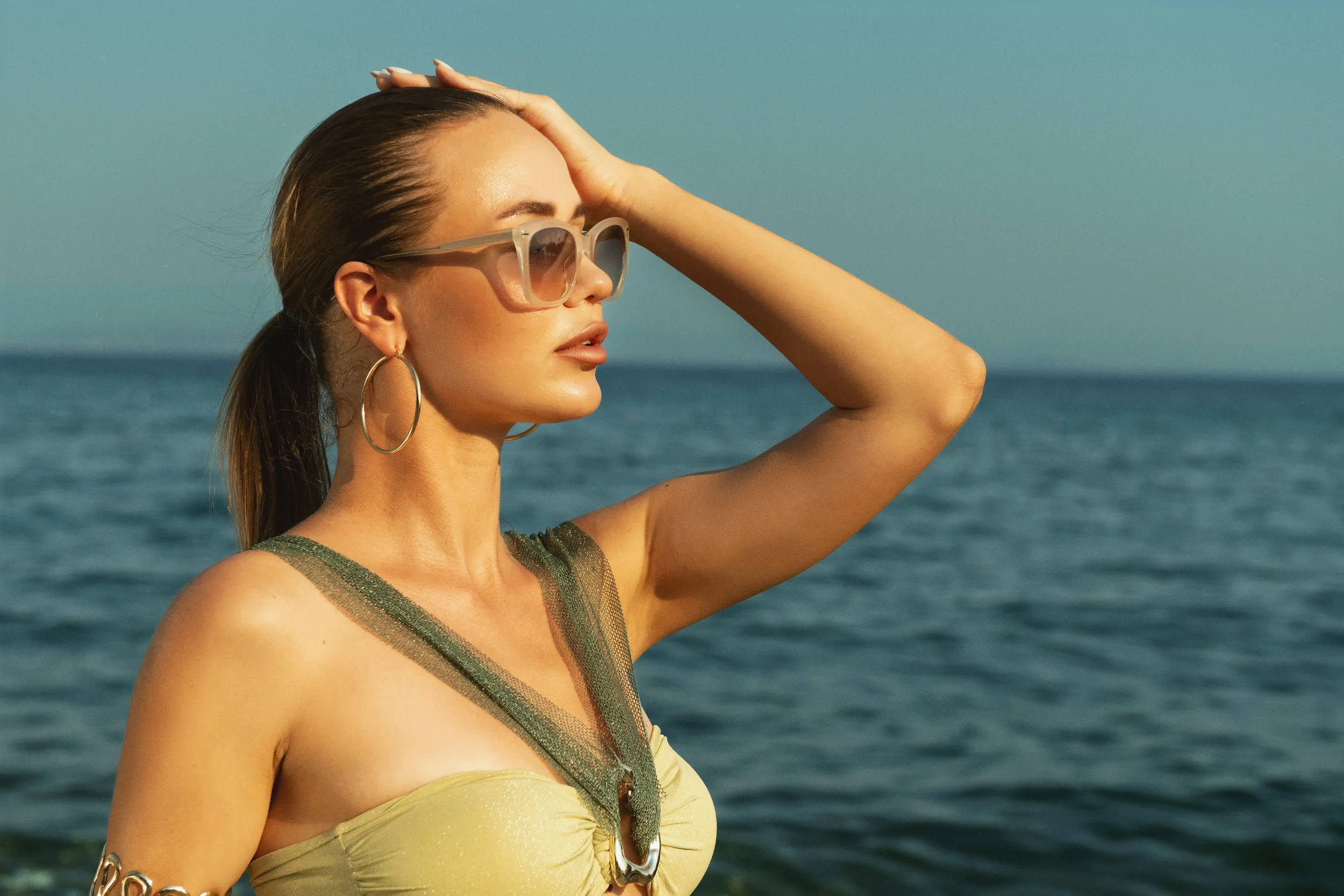 A woman with sunglasses and hoop earrings stands near the ocean, holding her hand to her head as she gazes into the distance in the sunlight.