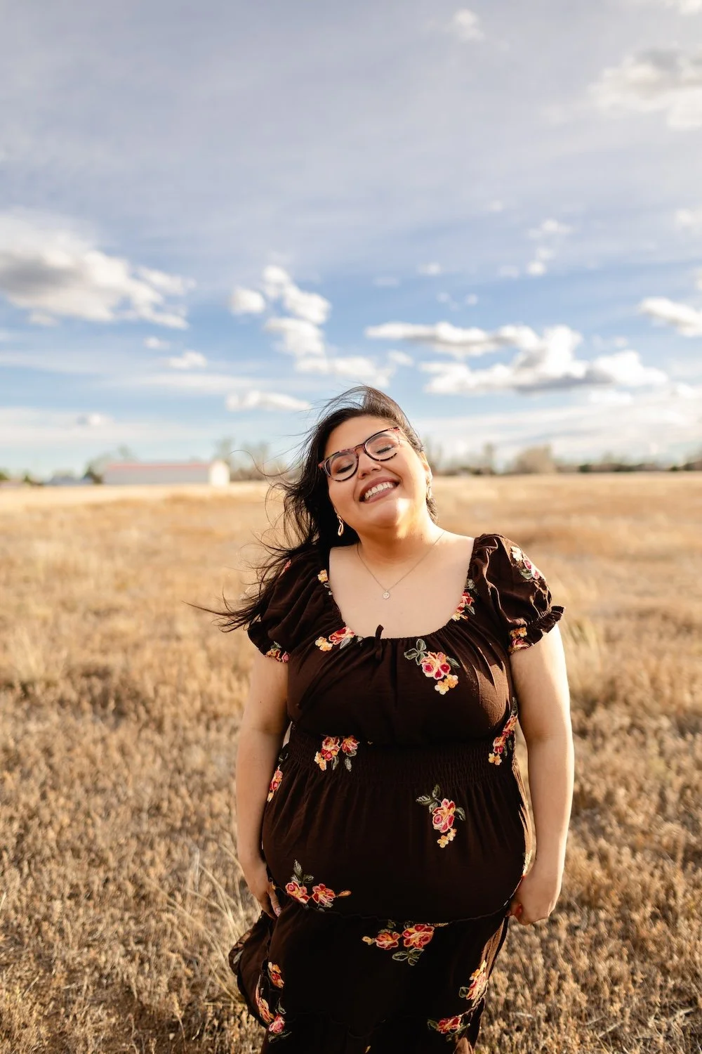 A woman smiling with glasses in a field under a blue sky with scattered clouds.