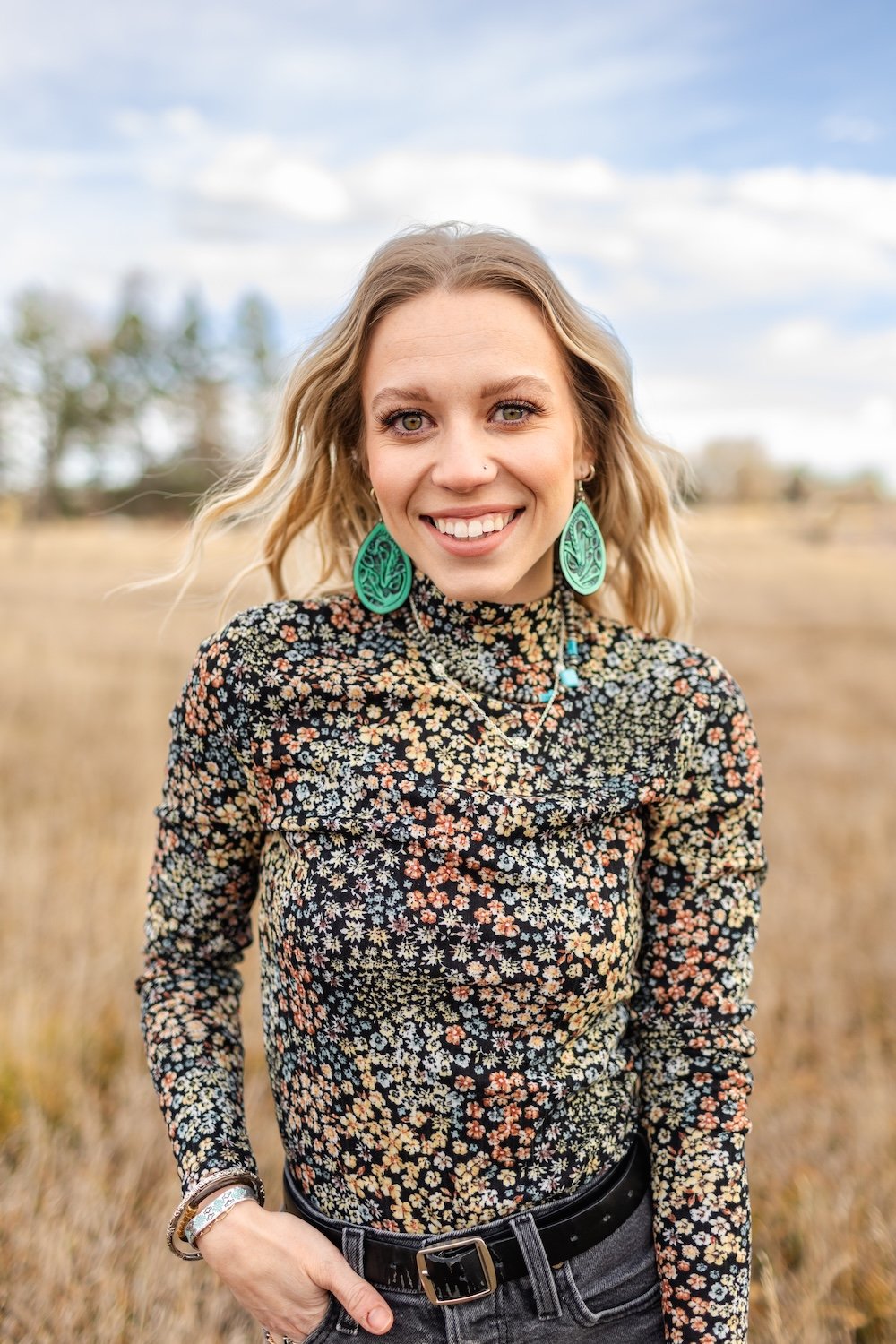 A smiling blonde woman with earrings and a floral top stands in a field with trees and a cloudy sky behind her.