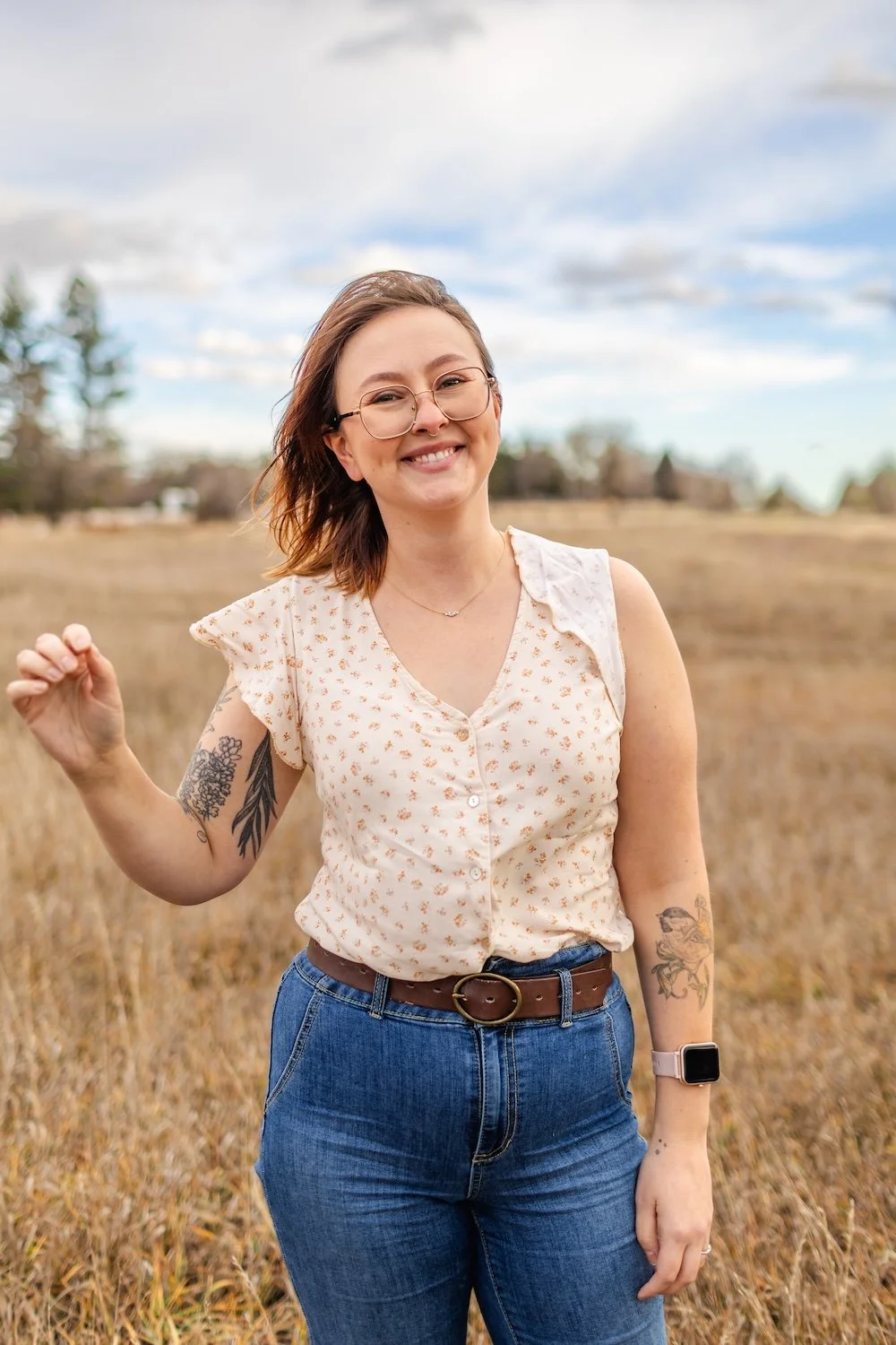A smiling woman with glasses standing in a field with tall grass under a partly cloudy sky.