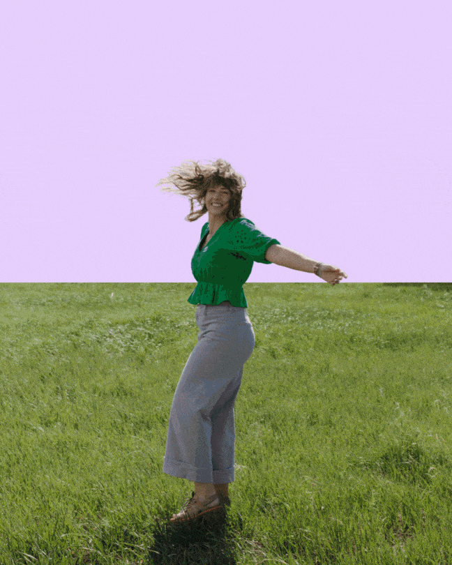 Woman with long curly hair dancing in a grassy field, wearing a green top and gray pants, with a purple sky background.