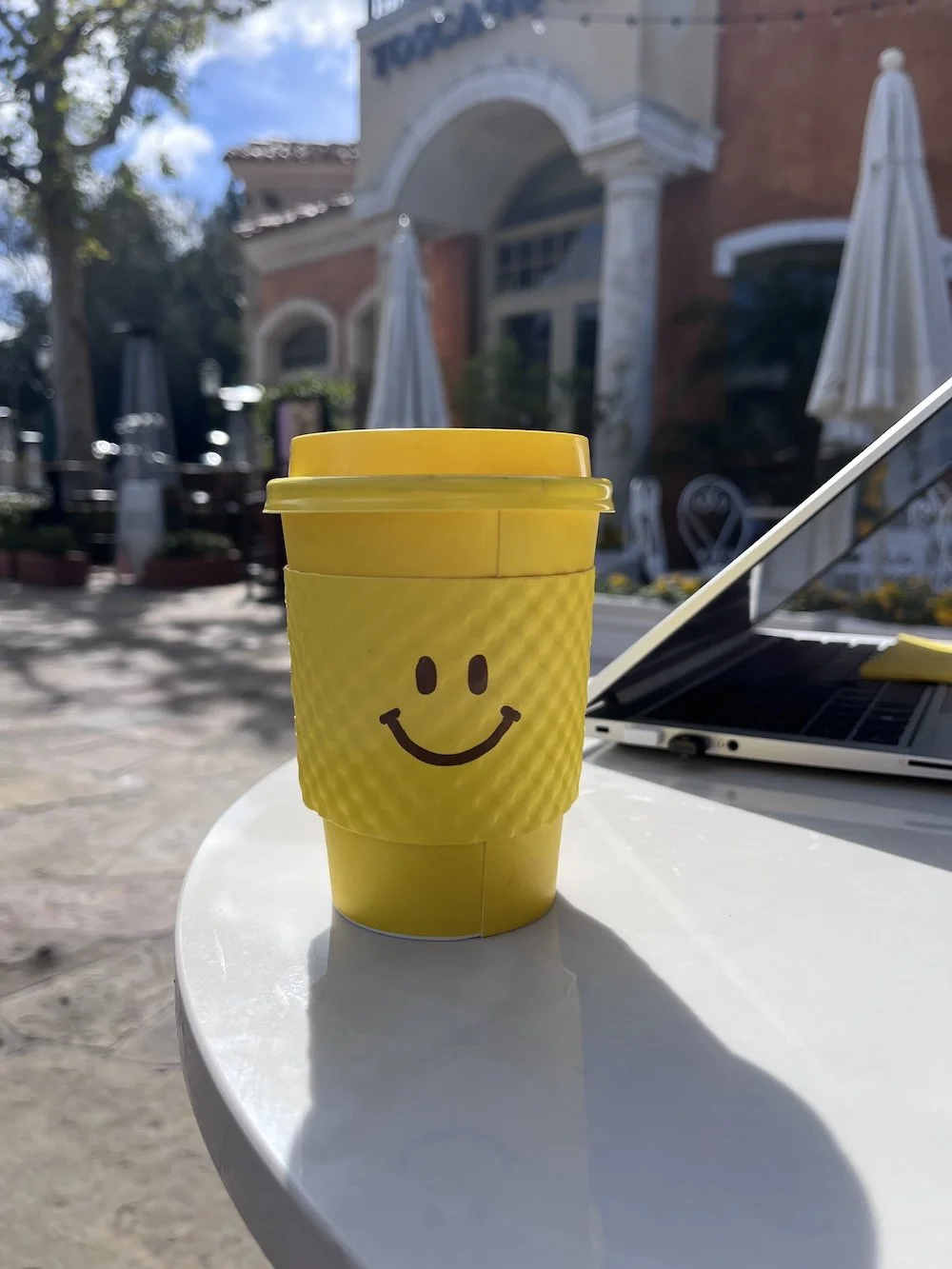 A yellow disposable coffee cup with a smiley face drawn on it, placed on a white table outdoors with umbrellas, trees, and a building in the background.