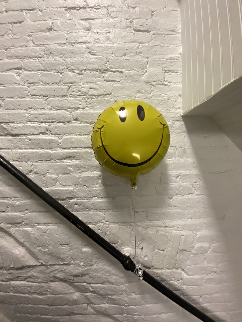 Yellow smiley face balloon attached to a white brick wall.