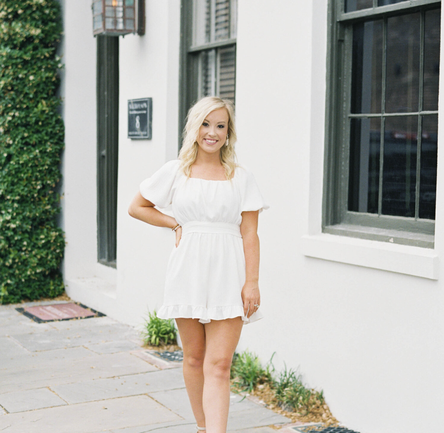 A woman in a white dress smiling while walking outside next to a white building with black window frames.