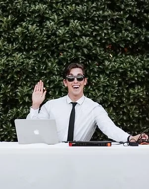 A woman in a white shirt and black tie, wearing sunglasses, sitting at a desk with a laptop and audio equipment, in front of a green leafy hedge, smiling and waving.