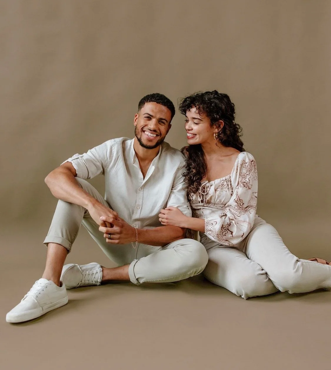 A smiling couple sitting on the floor against a beige background, with the woman leaning on the man.
