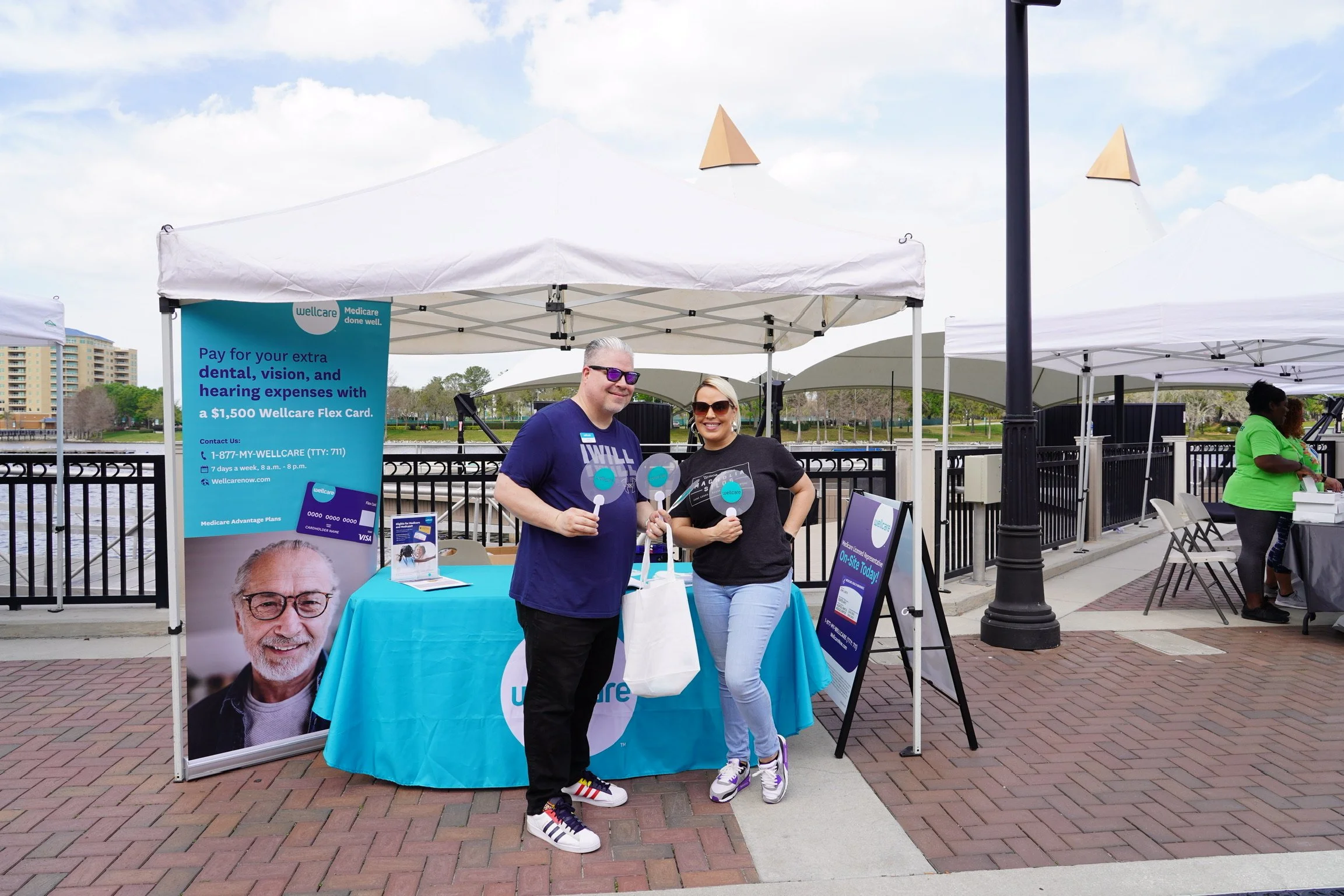Two people standing under a white canopy tent at an outdoor event, holding promotional signs for Wellcare. There is a large blue banner advertising dental, vision, and hearing plans, with a picture of a smiling older man. They are on a brick walkway 