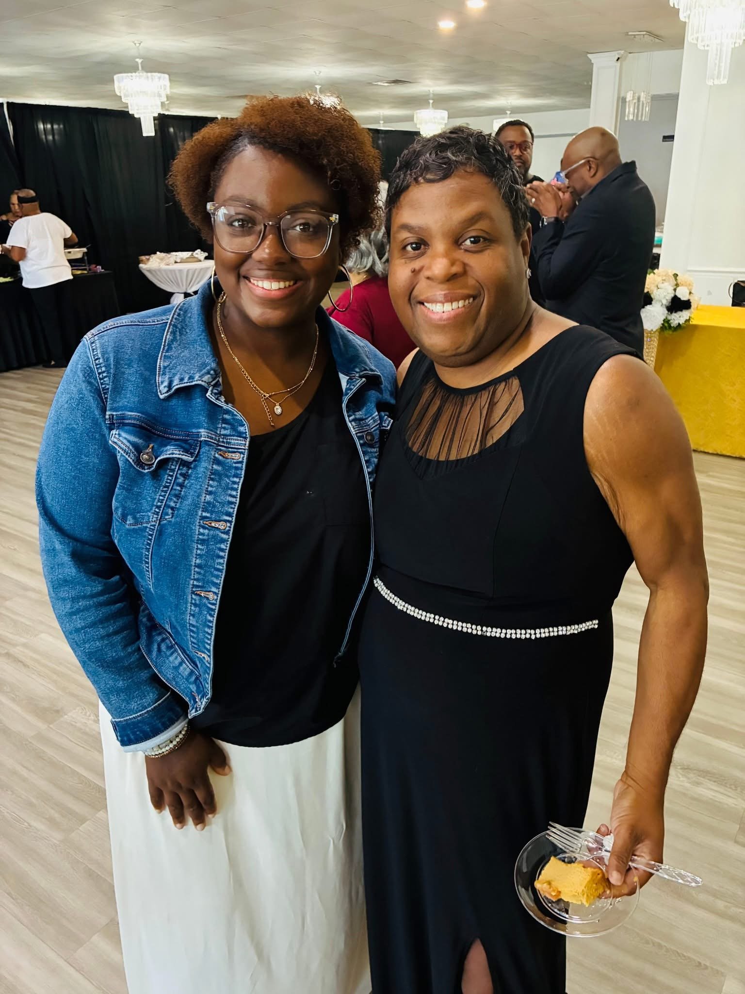 Two women smiling at a social event, one wearing a denim jacket and glasses, the other in a black dress holding a plate with a piece of cake.
