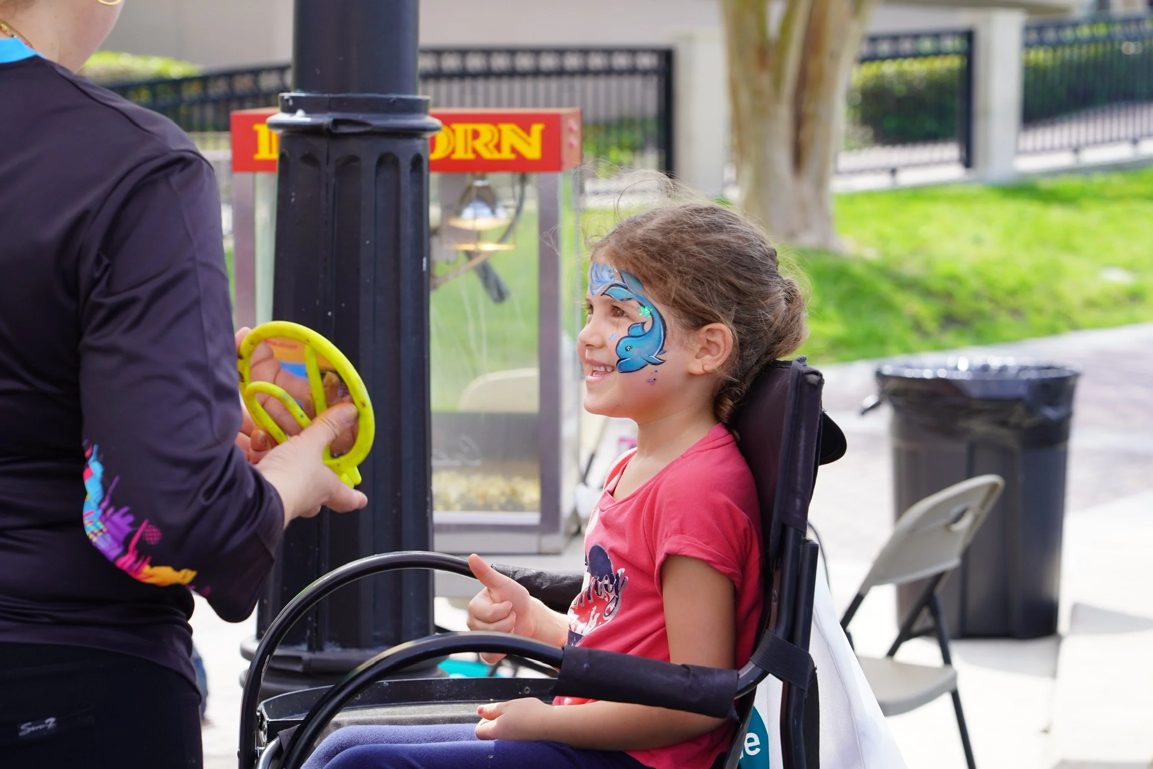 A young girl with a blue face painting of a mermaid on her cheek, sitting in a wheelchair, smiling as a woman holds a yellow bubble wand near her. There is popcorn stall in the background and a trash bin nearby.