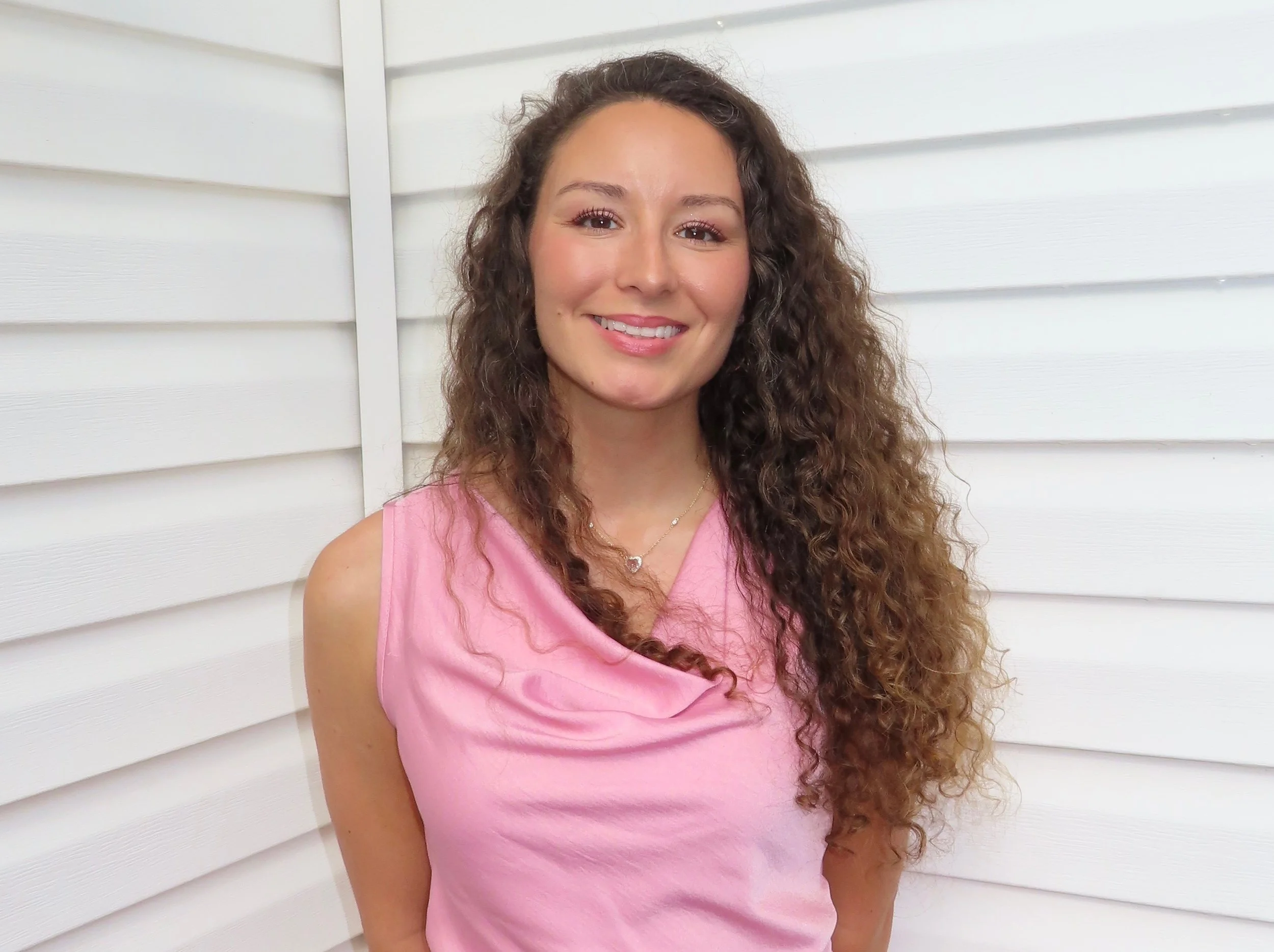 A woman with long, curly brown hair smiling, wearing a pink sleeveless top and a heart-shaped necklace, standing against a white wooden wall.