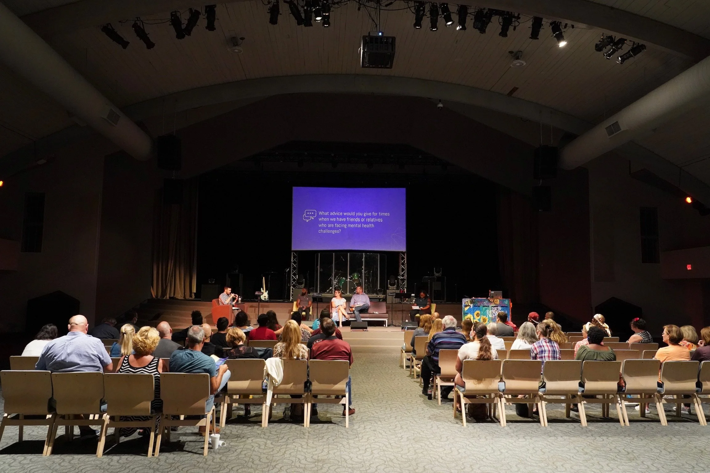 A panel discussion taking place on a stage in a large auditorium with a seated audience, a screen displaying a question about mental health advice, musical instruments on stage, and a painted piano on the right.