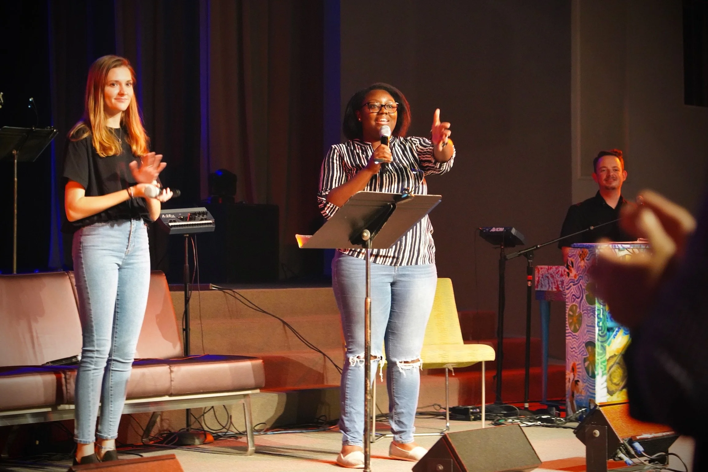 Three people on stage during a performance or speech. The woman in the center is speaking into a microphone, wearing glasses, a striped shirt, and ripped jeans. To her left, a young woman is holding a microphone with a hand raised, wearing a black t-shirt and light blue jeans. To the right, a man is playing a keyboard, dressed in black, smiling. There are musical instruments and chairs on stage, with some audience members visible.