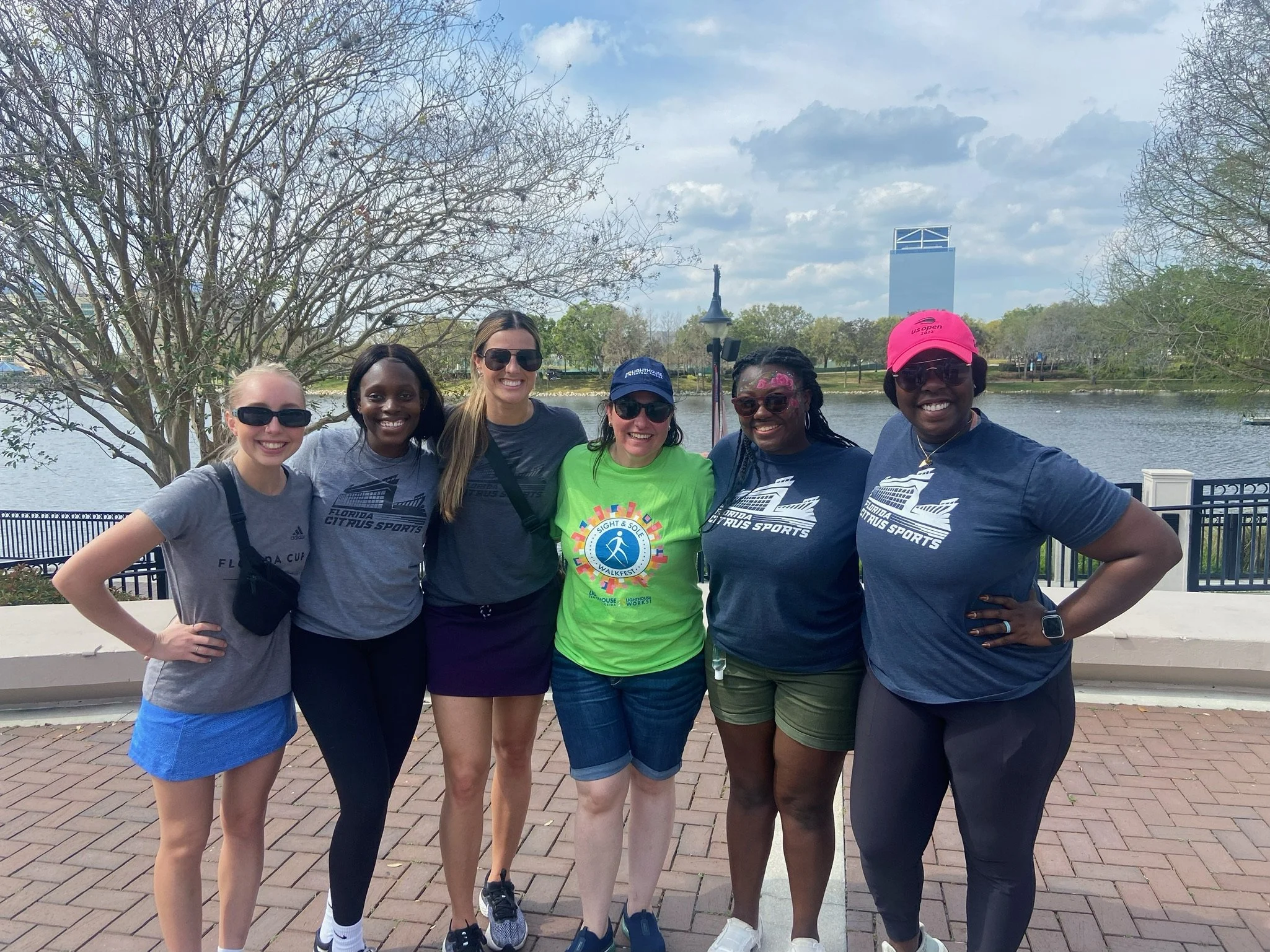 Group of six women standing outdoors near a lake, smiling.