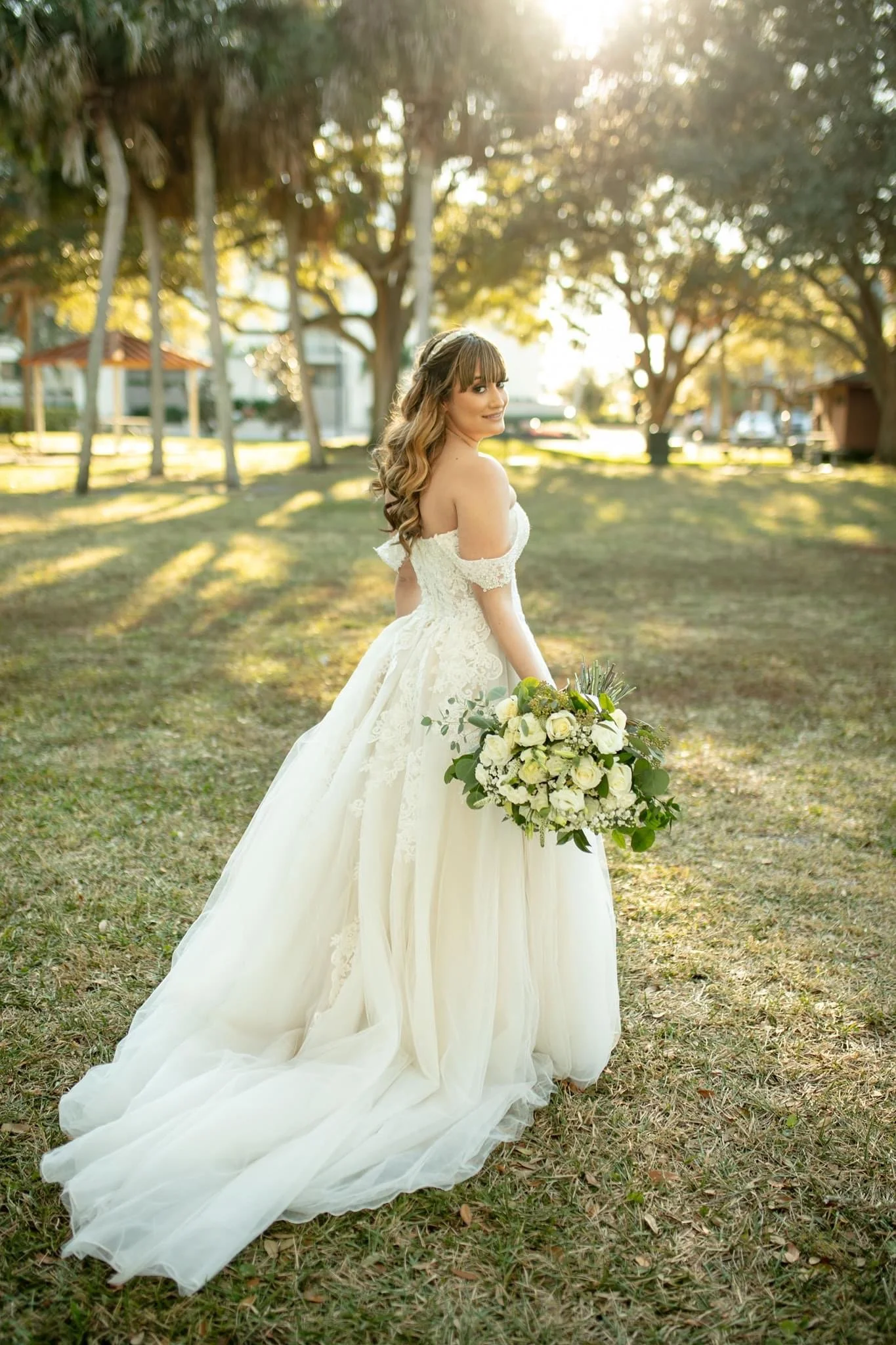 A bride in a white wedding gown holding a bouquet of white roses and greenery, standing outdoors on grass with trees and sunlight in the background.