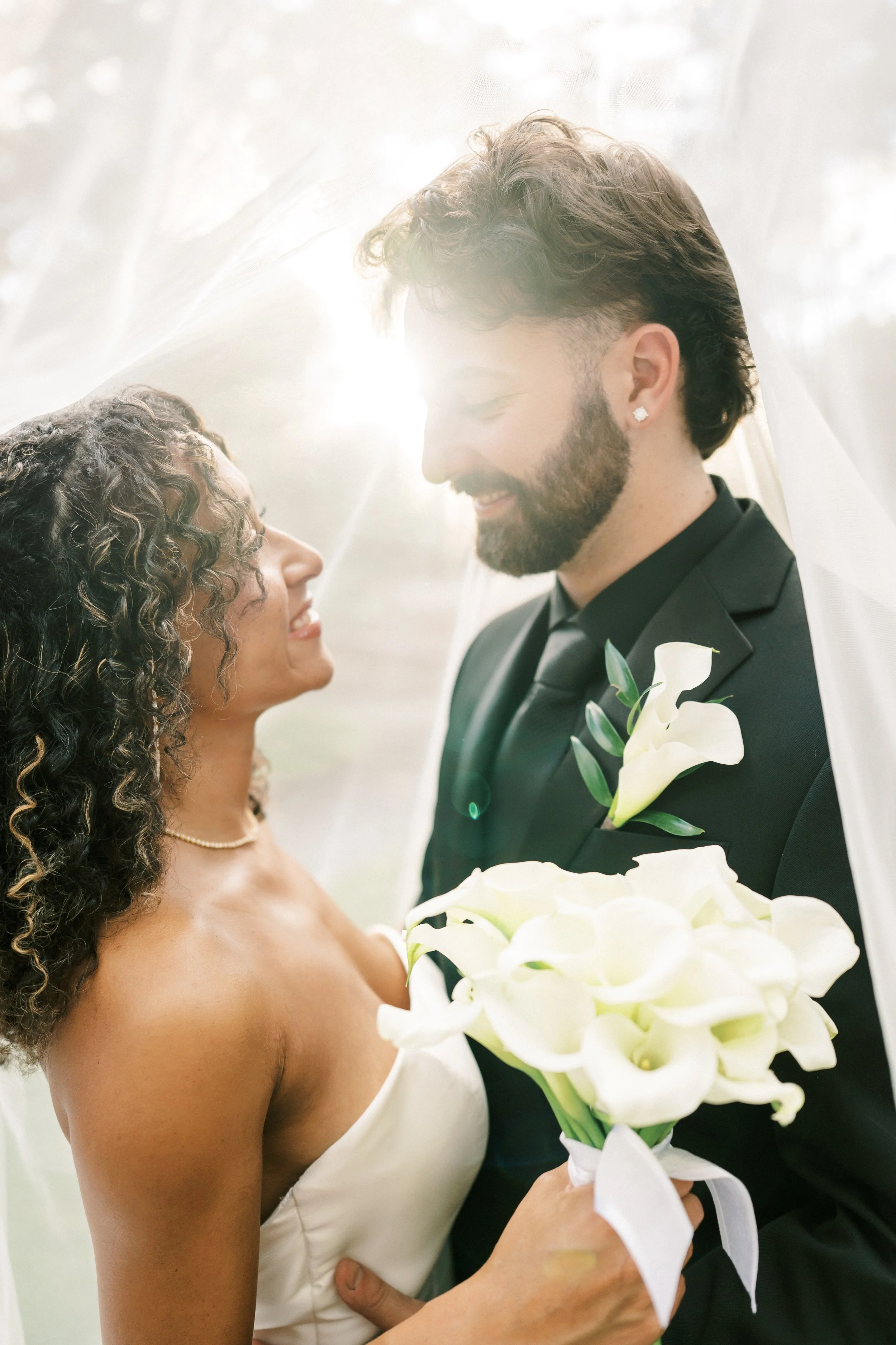 A bride and groom smiling at each other, with a veil and bouquet of white calla lilies, on their wedding day.