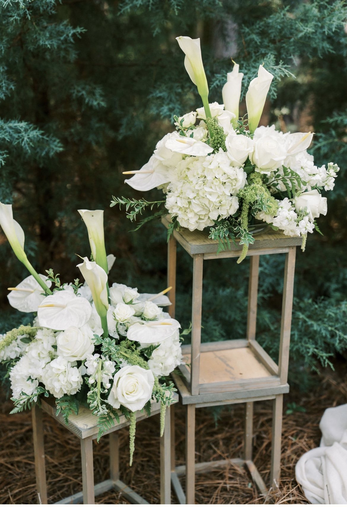 Two floral arrangements of white flowers, including roses, hydrangeas, lilies, and calla lilies, placed on wooden stands, against a backdrop of greenery.