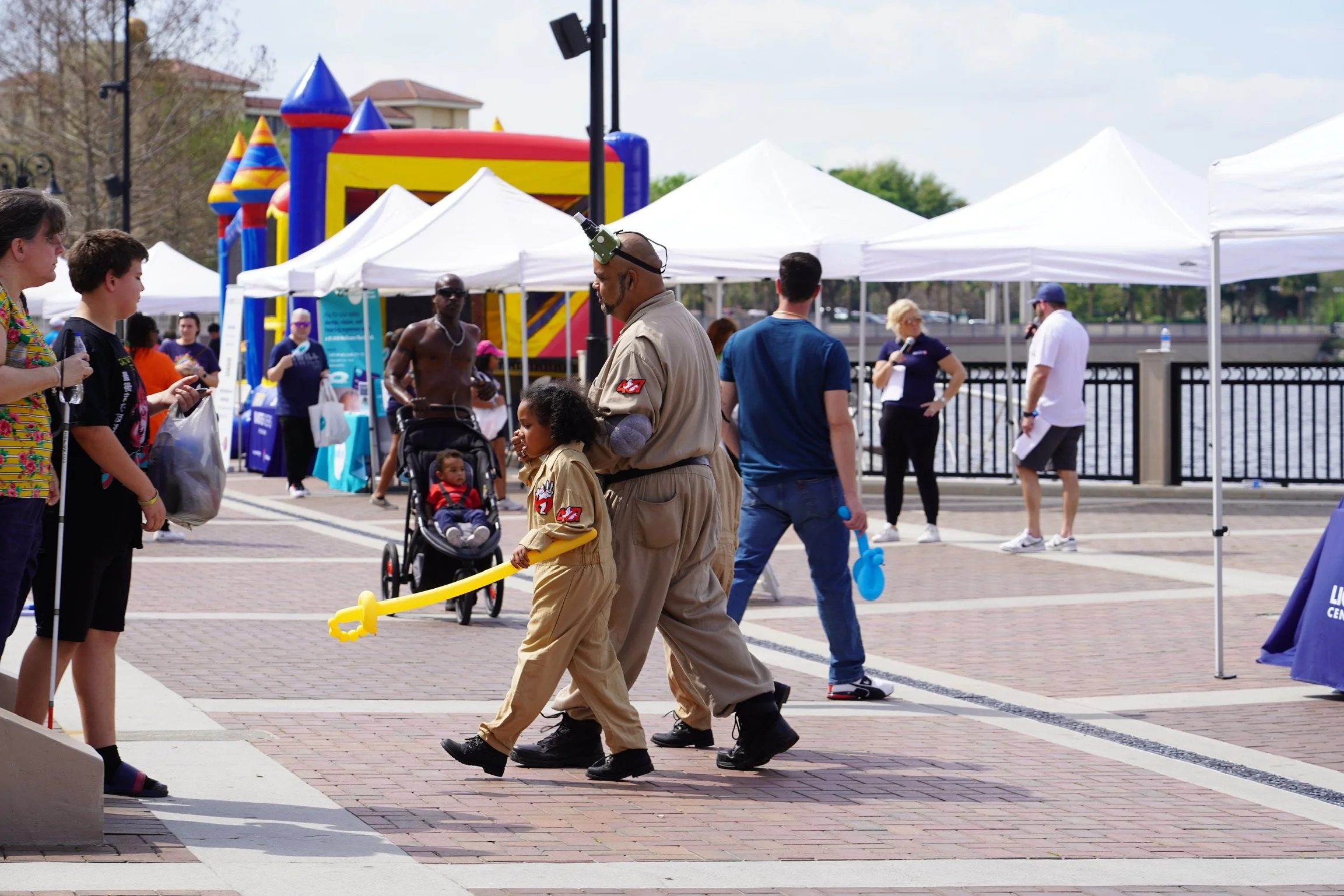 A scene at an outdoor event with various people, booths, and a bounce house in the background. A man and a young girl, both dressed in beige uniforms, walk together, with the girl holding a yellow toy sword. Other people are socializing, walking, or 