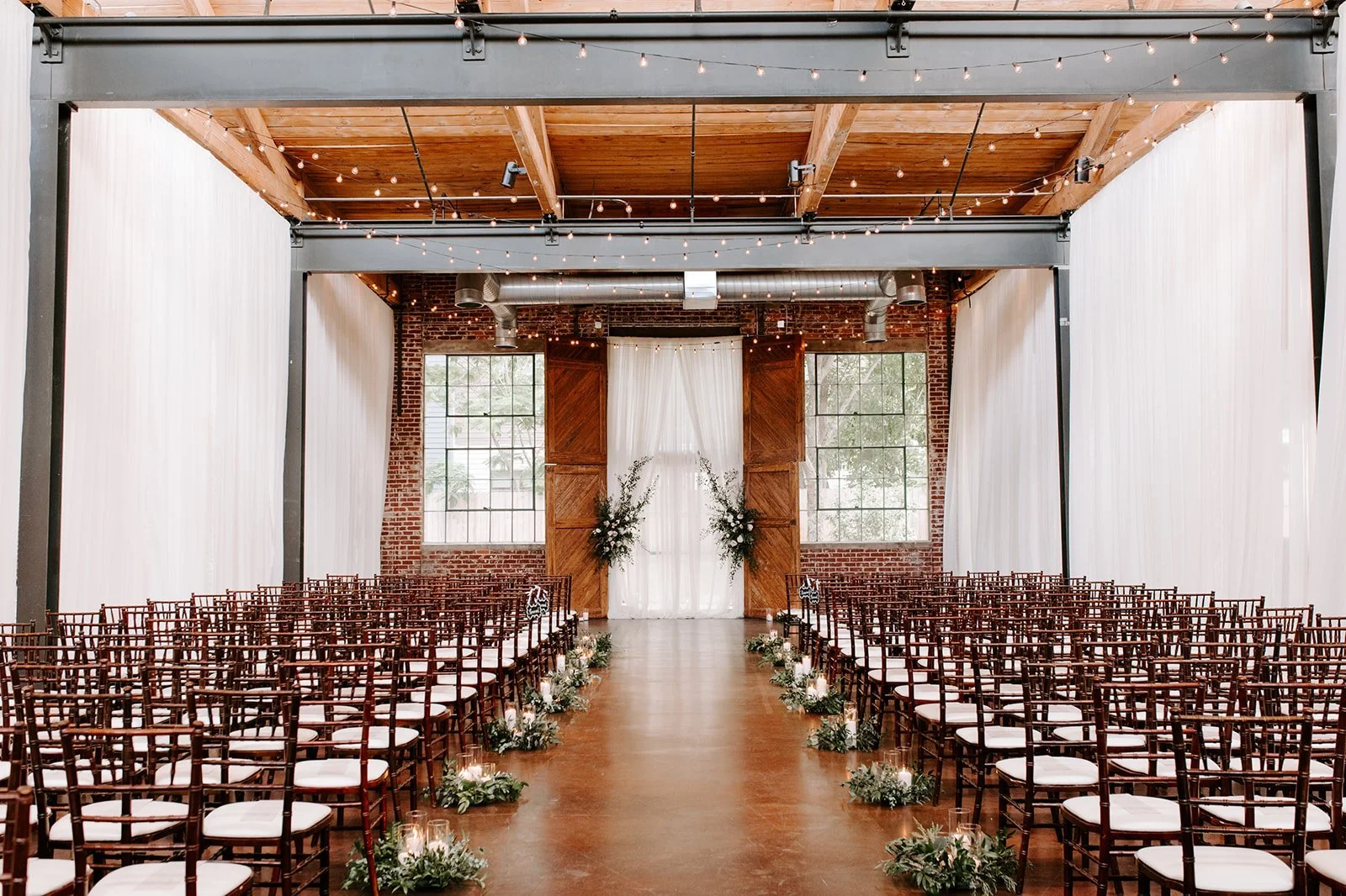 Indoor wedding ceremony setup with rows of wooden chairs with white cushions, floral arrangements on the aisle, a white curtain backdrop, and large industrial-style windows.