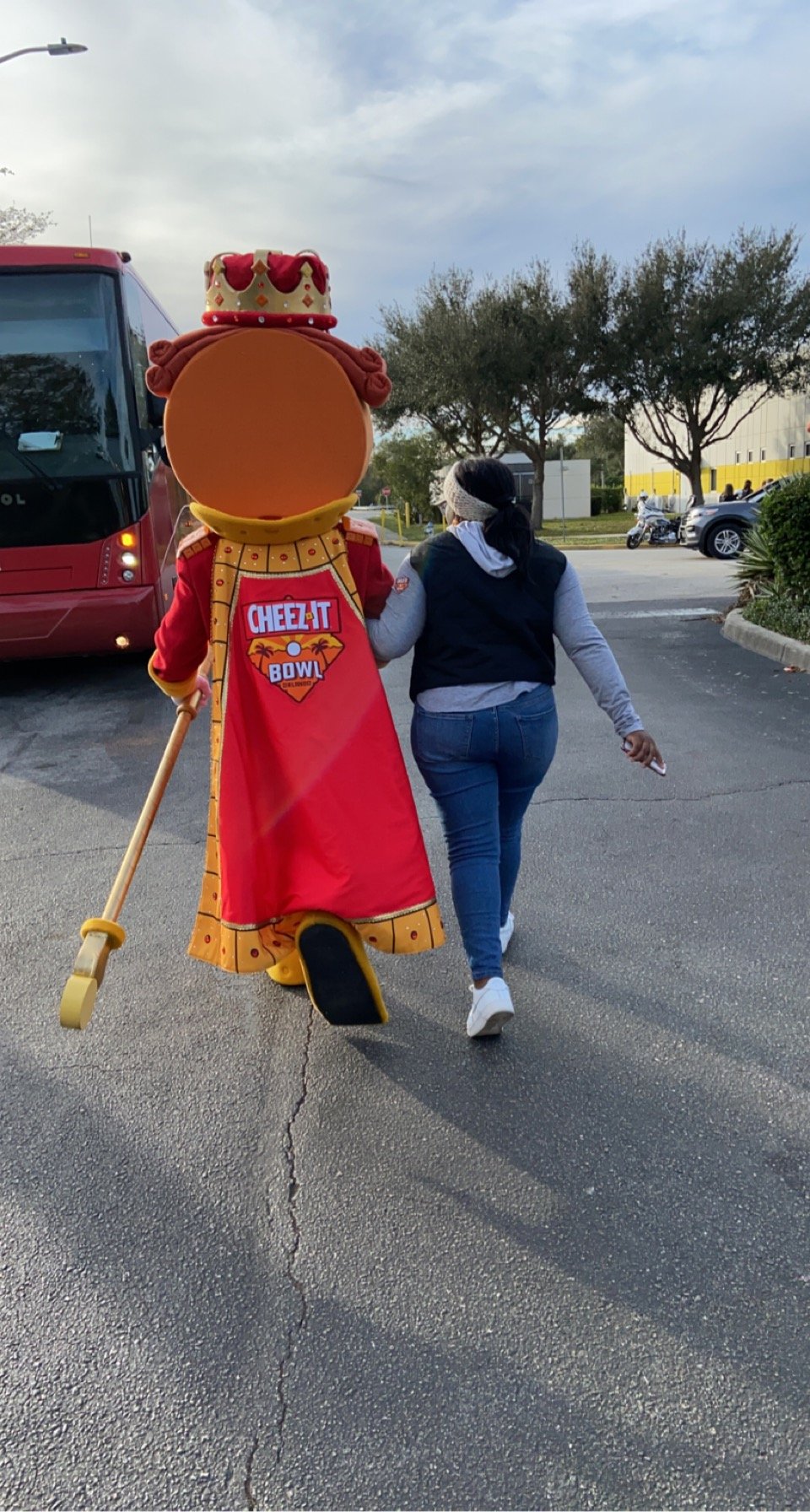 A woman walking with a person dressed in a Costumed mascot of a character in a Cheerleader outfit with a crown, red cape, and yellow scepter, in a parking lot with trees and cars in the background.