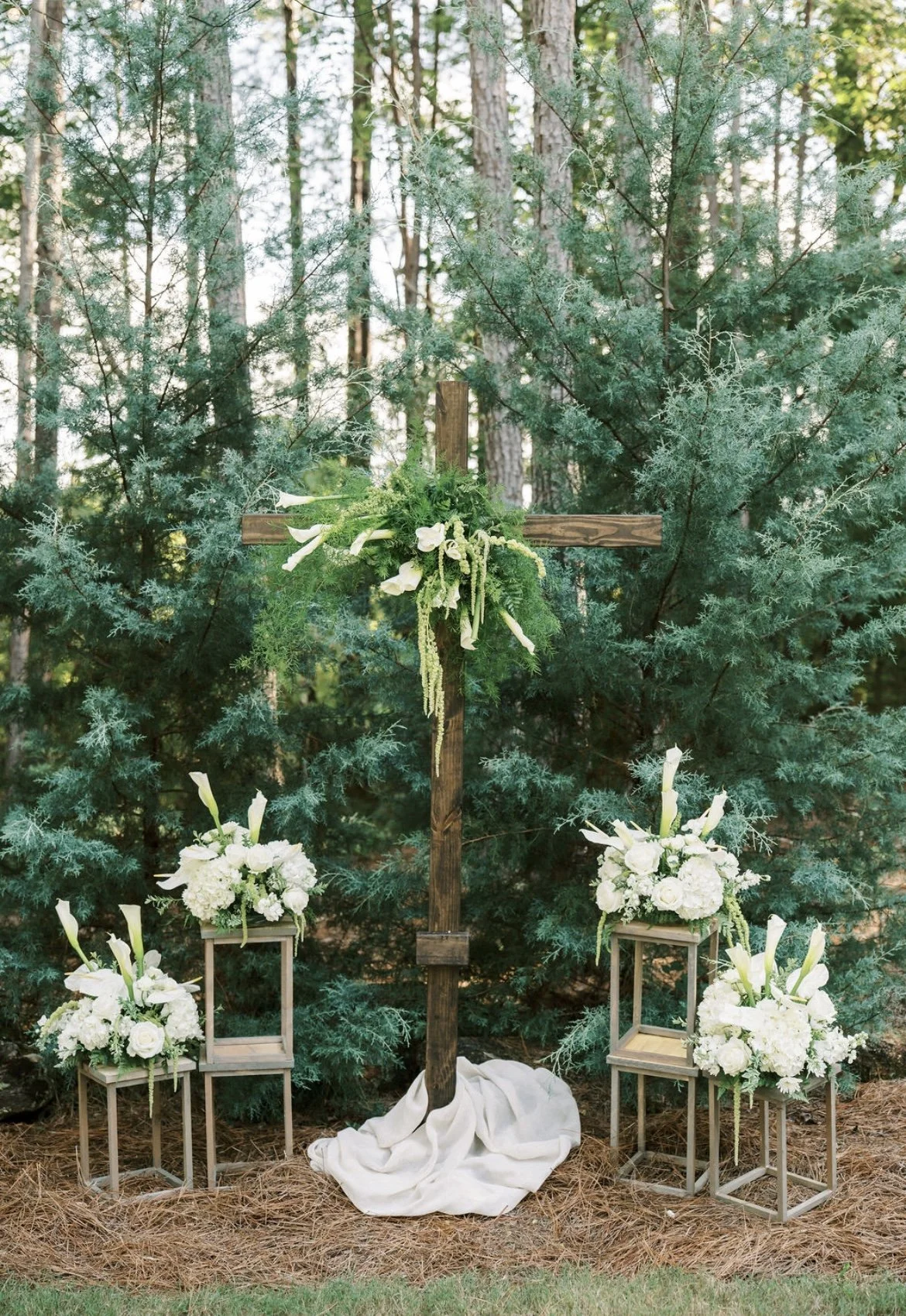 A wooden cross decorated with green foliage and white flowers stands outdoors against green pine trees. There is a white cloth draped at the base of the cross. Two arrangements of white flowers are placed on small beige stools on either side of the c