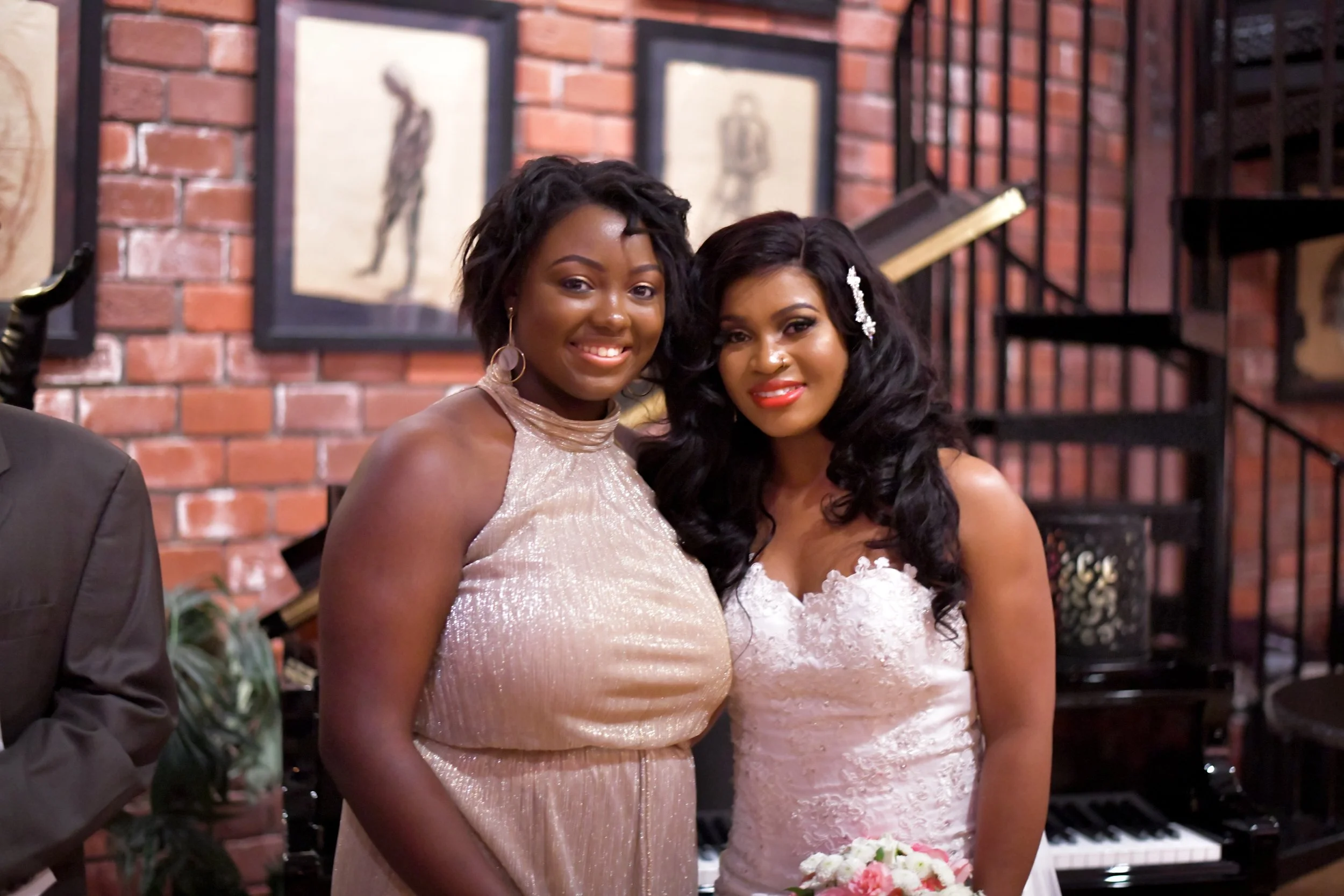 Two women in formal dresses smiling at an indoor event with brick walls and artwork in the background.