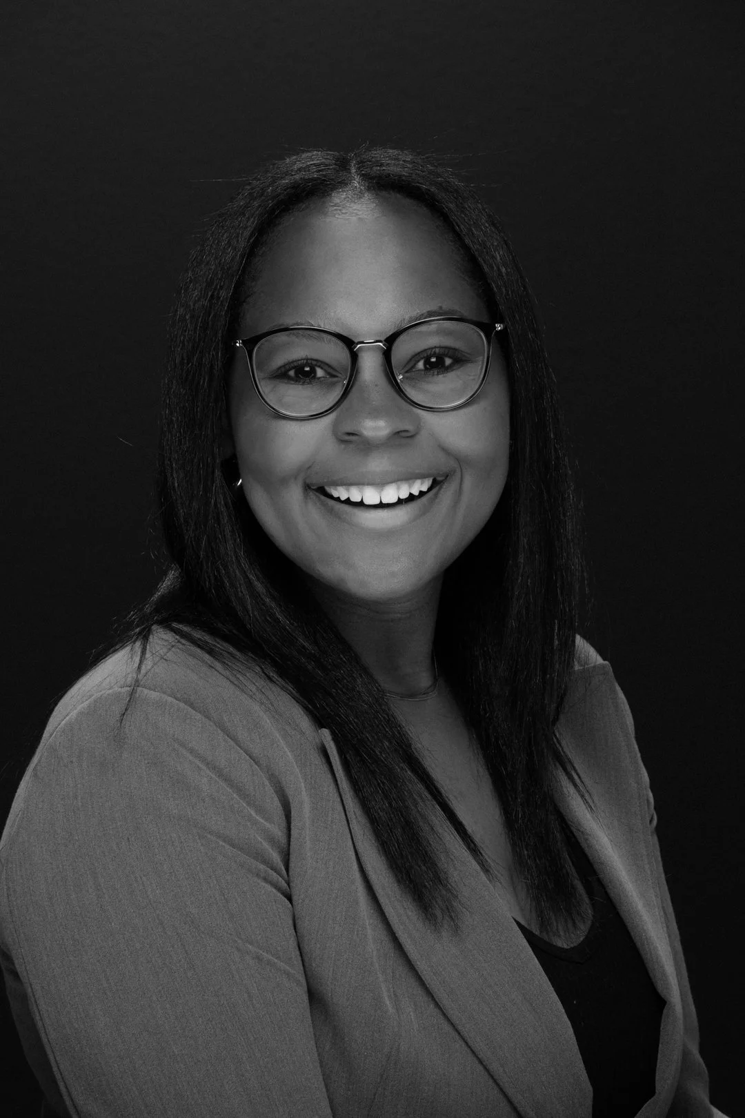 A smiling woman with glasses, long dark hair, and a blazer against a dark background.