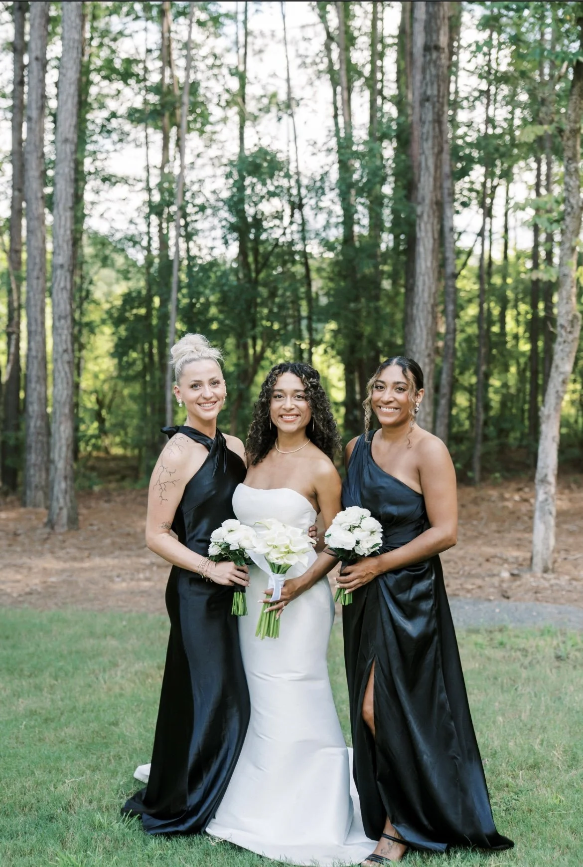 Three women standing outdoors in a wooded area, dressed for a wedding. The woman in the center is wearing a white strapless wedding gown and holding a bouquet of white flowers. The other two women are wearing black dresses and holding matching bouque