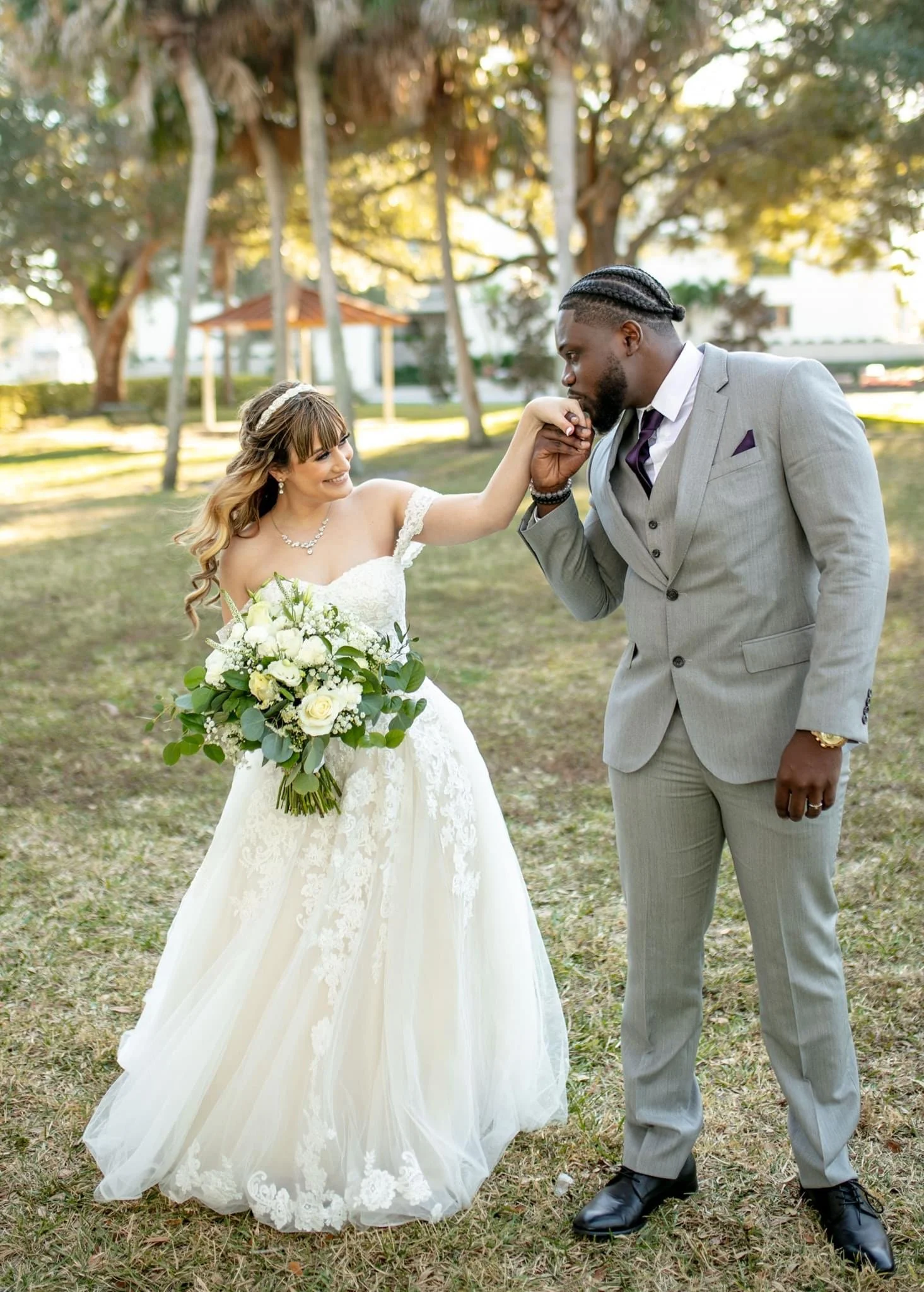 A bride and groom in a park, with the groom kissing the bride's hand. The bride is smiling and holding a bouquet of white roses and greenery, wearing a white wedding dress. The groom is dressed in a light gray suit with a purple tie, standing on grass with trees in the background.