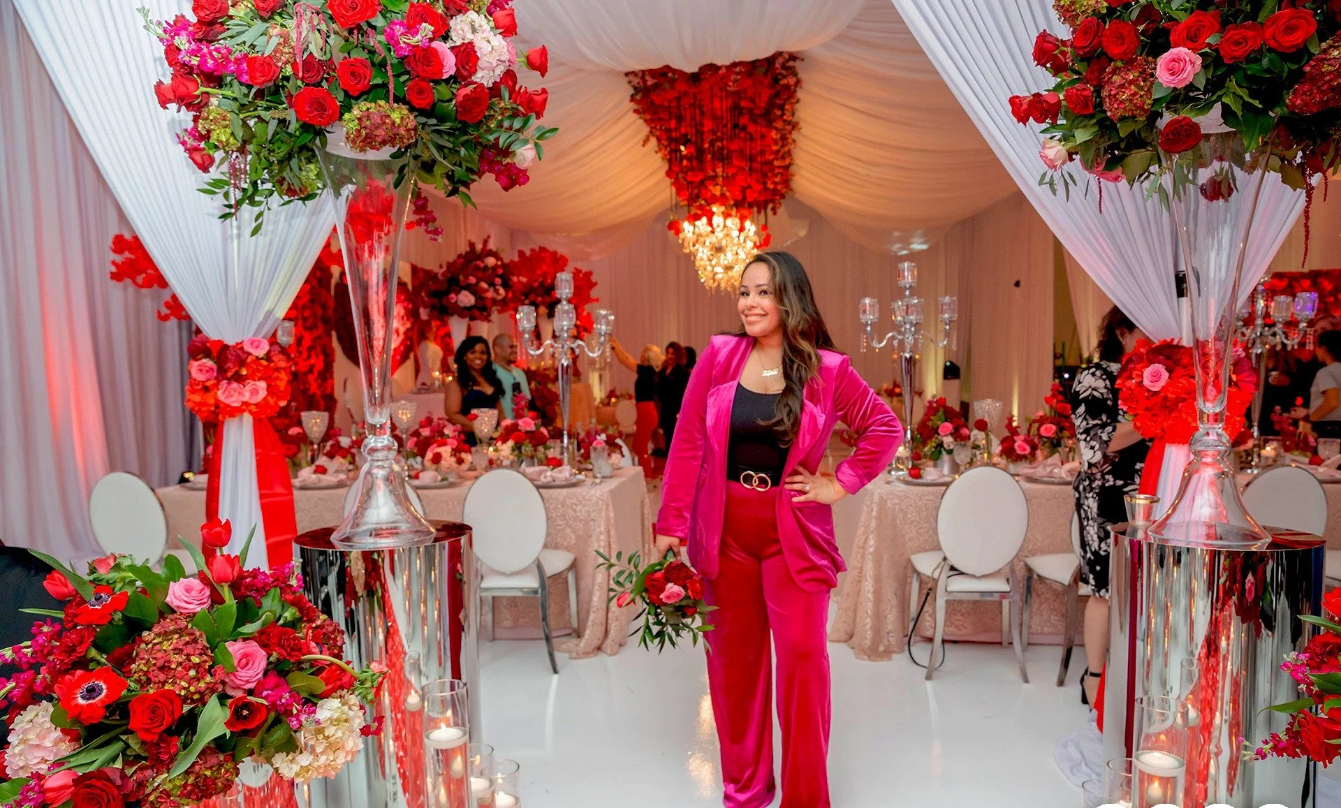 A woman in a bright pink suit smiling at a decorated event hall with red and pink floral arrangements, draped white fabric, and elegant table settings.