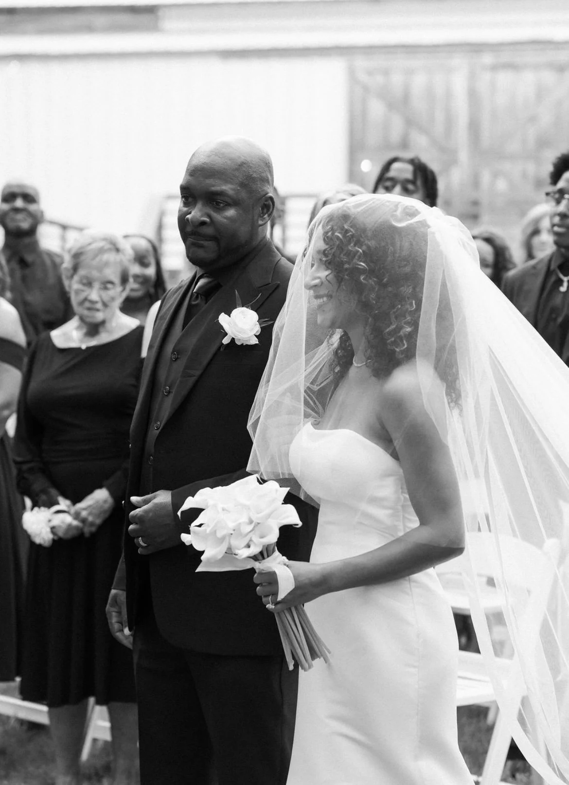 A bride in a strapless wedding dress with a veil holding a bouquet of calla lilies, standing next to a groom in a dark suit with a white flower boutonniere, during a wedding ceremony.