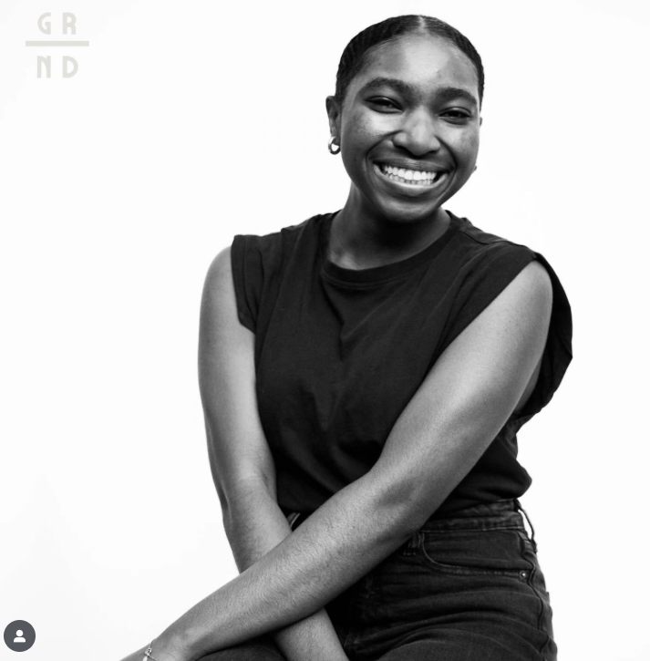 Black woman smiling, wearing a black sleeveless top and earrings, seated against a plain background.