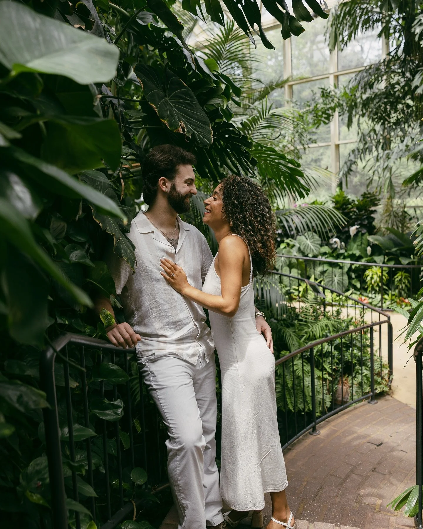A couple in white outfits sharing a joyful moment in a lush, green greenhouse with large tropical plants.