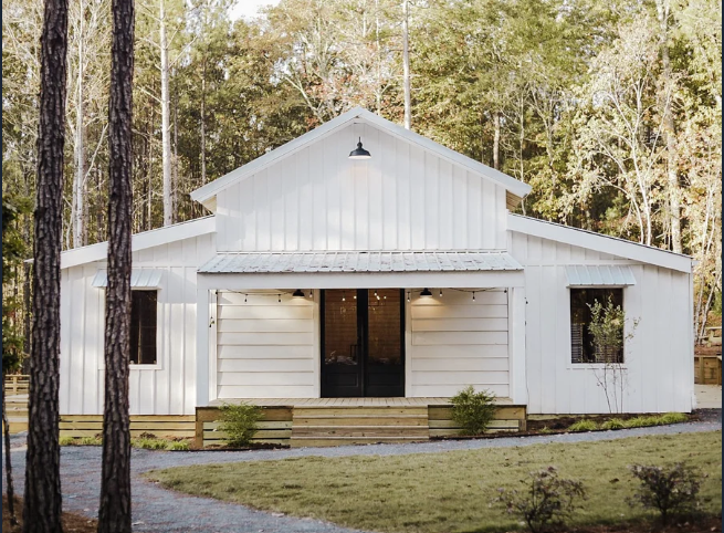 A white barn-style house with a front porch, surrounded by a grassy yard and woodland.