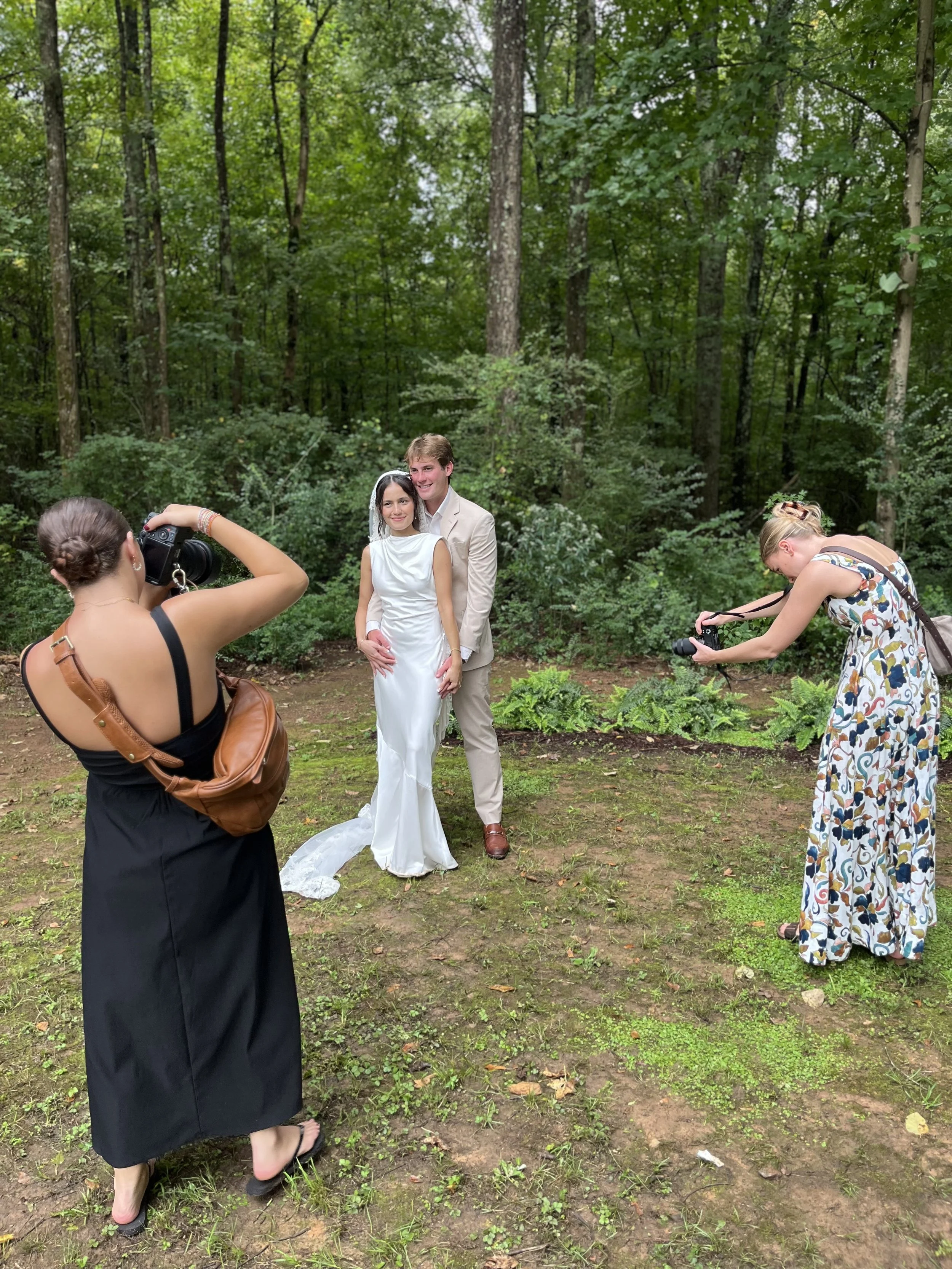 A bride and groom posing for photos in a wooded outdoor setting, with two women taking pictures of them.