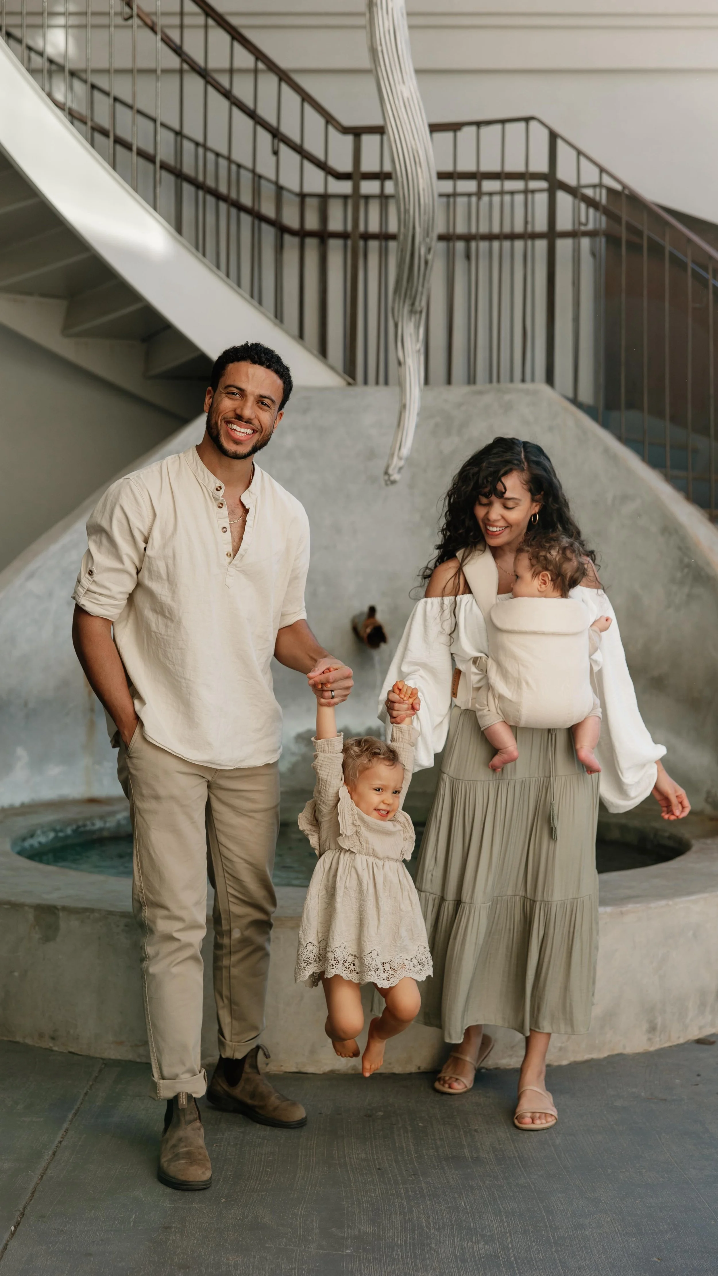 Family of four, smiling, holding hands, standing in front of water fountain inside a building with staircase and railing, casual beige and cream clothing, baby in carrier, daughter jumping, modern interior.