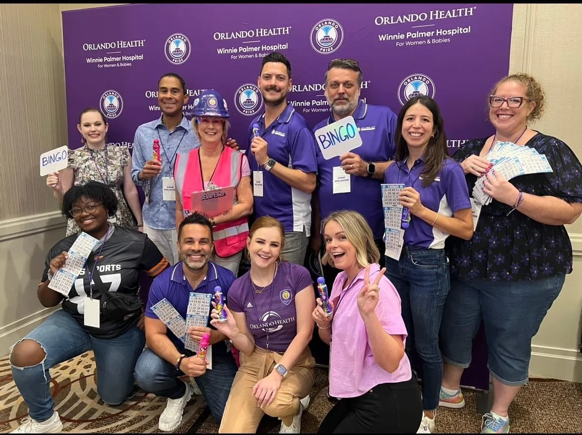 Group of people smiling and holding bingo tickets and prizes at a bingo event hosted by Orlando Health at Winnie Palmer Hospital. The backdrop displays Orlando Health branding.