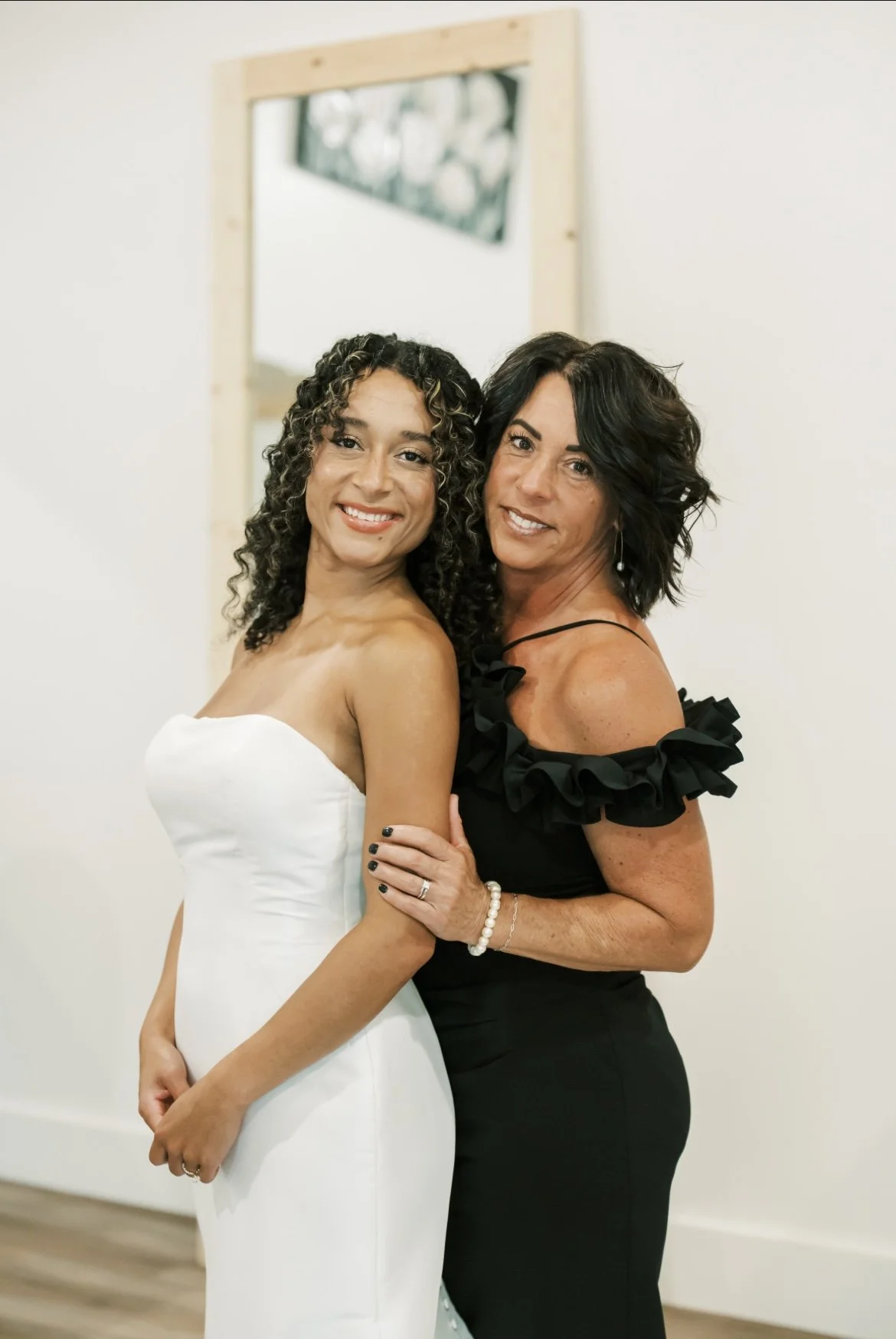 Two women standing close together, smiling at the camera. The woman on the left is wearing a white dress, and the woman on the right is dressed in black with ruffled sleeves. They are indoors, with a mirror and part of a window visible in the backgro