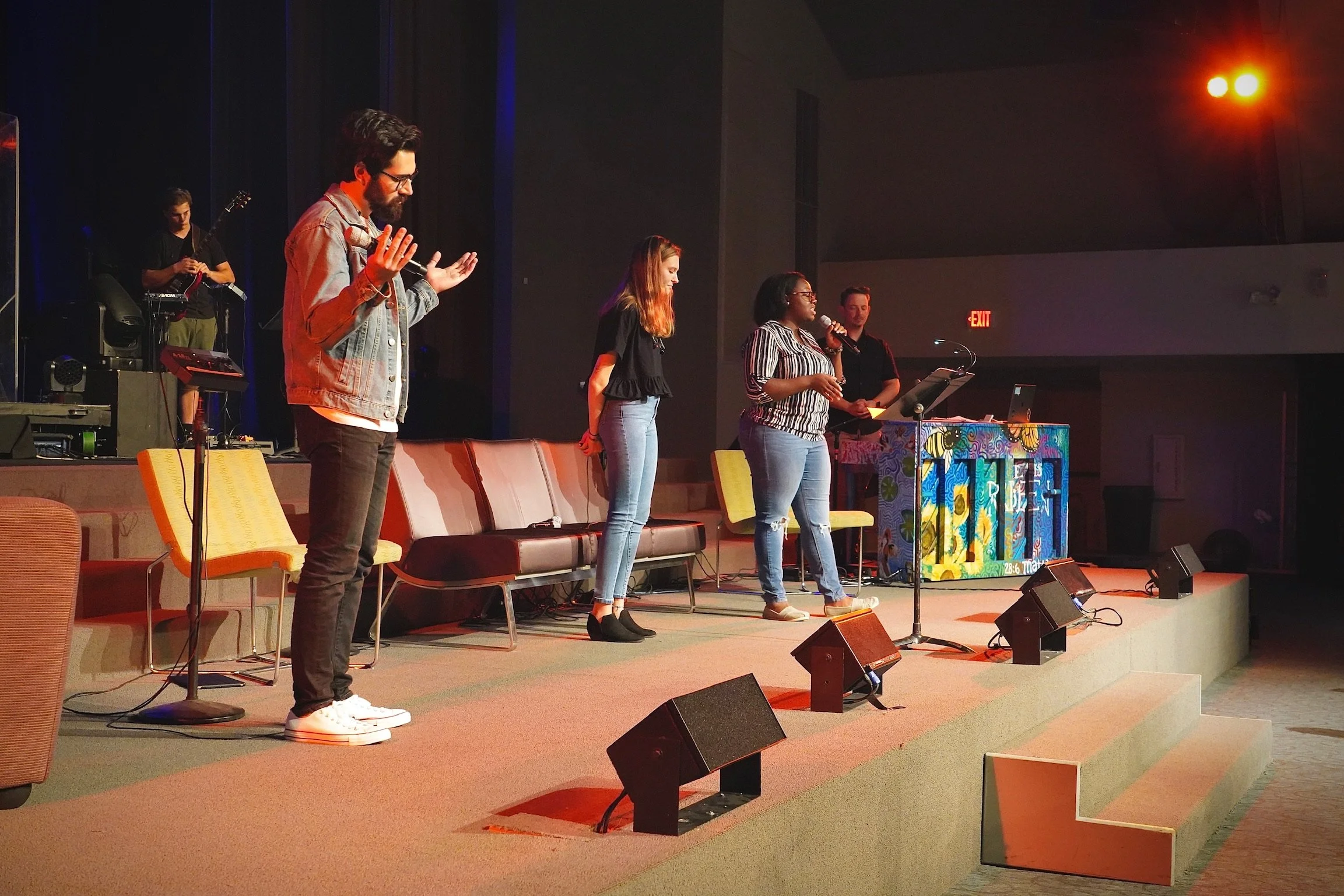 Group of four people on stage during a musical or worship event, with one person playing guitar in the background and the remaining three singing or speaking into microphones, with colorful stage lighting and an acoustic setup.