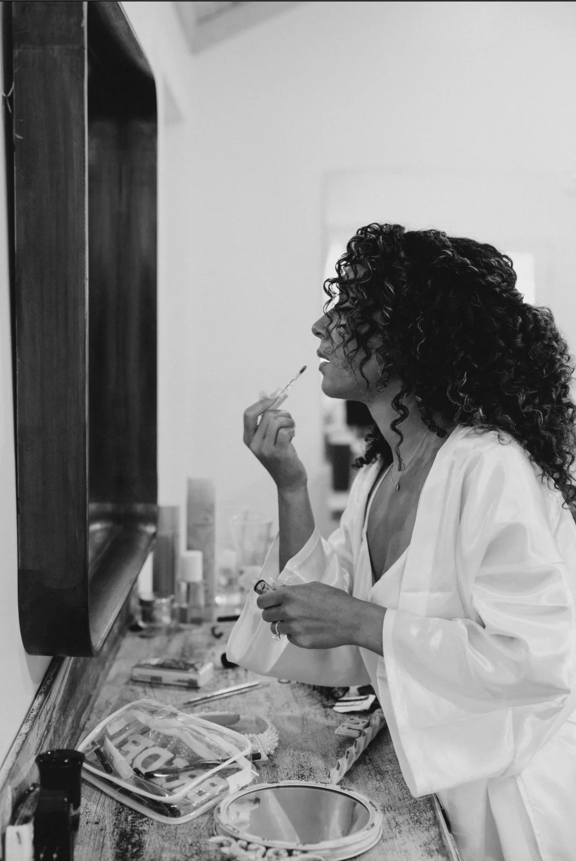 A woman with curly hair in a white robe applies lipstick in front of a mirror in a vanity or dressing room.