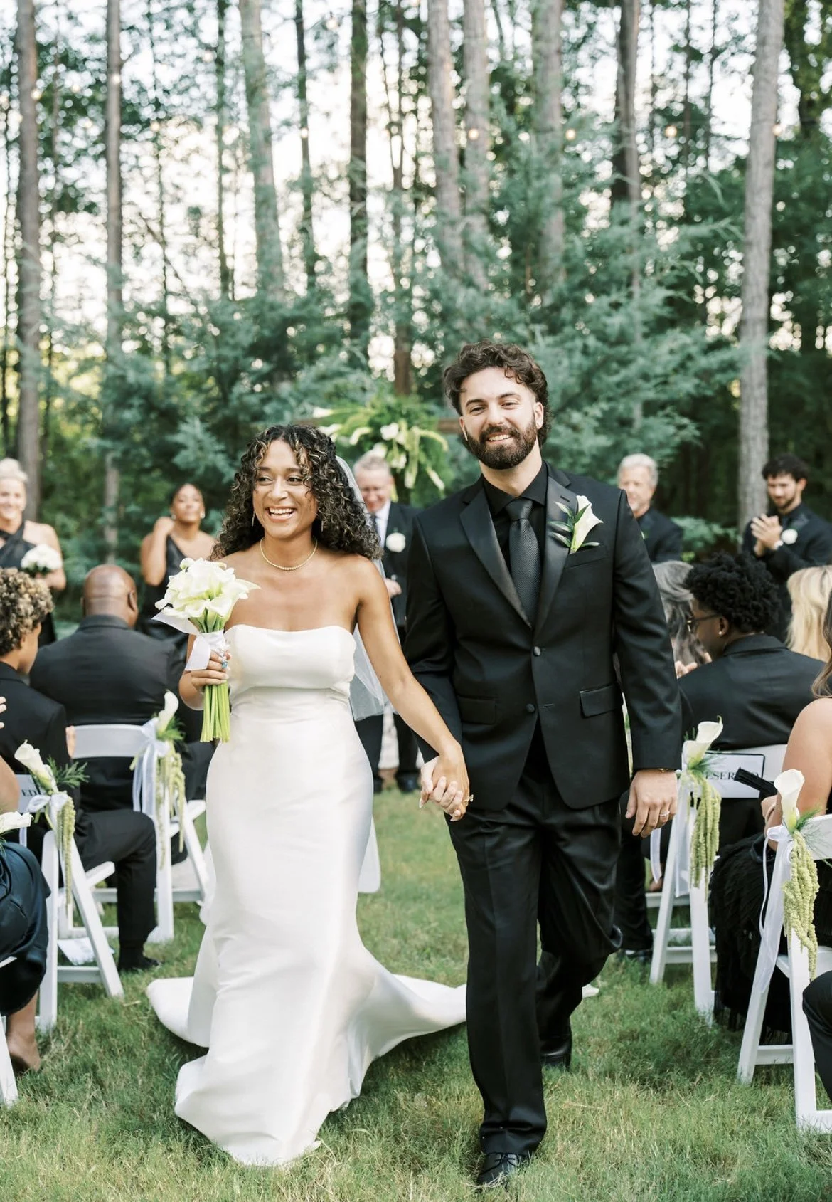 A bride and groom walking hand in hand at their outdoor wedding ceremony, surrounded by seated guests in a forested area.