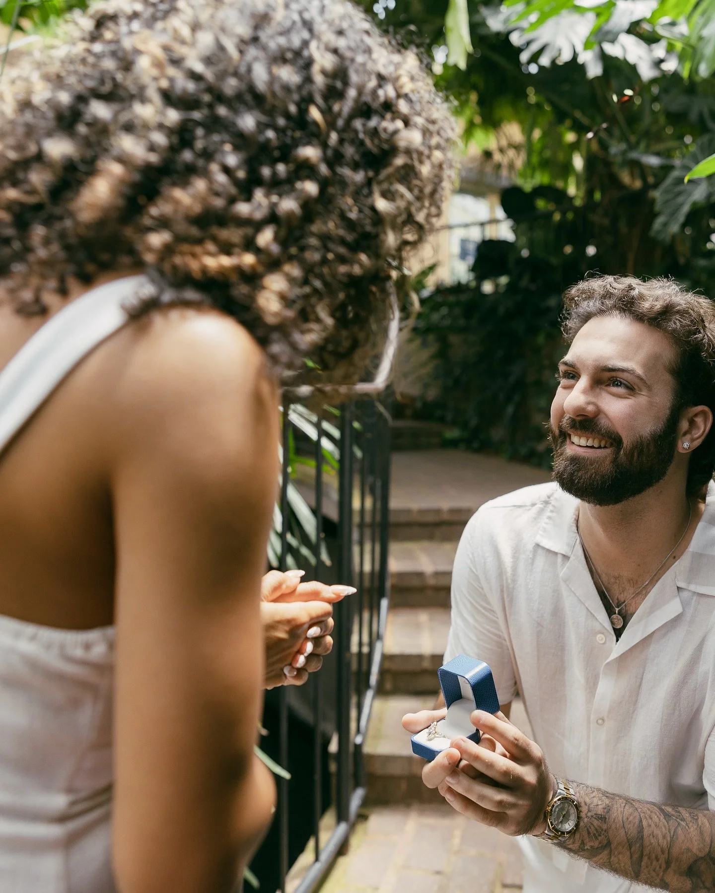 A man is proposing marriage to a woman outdoors, holding an open ring box, while the woman reacts with excitement.