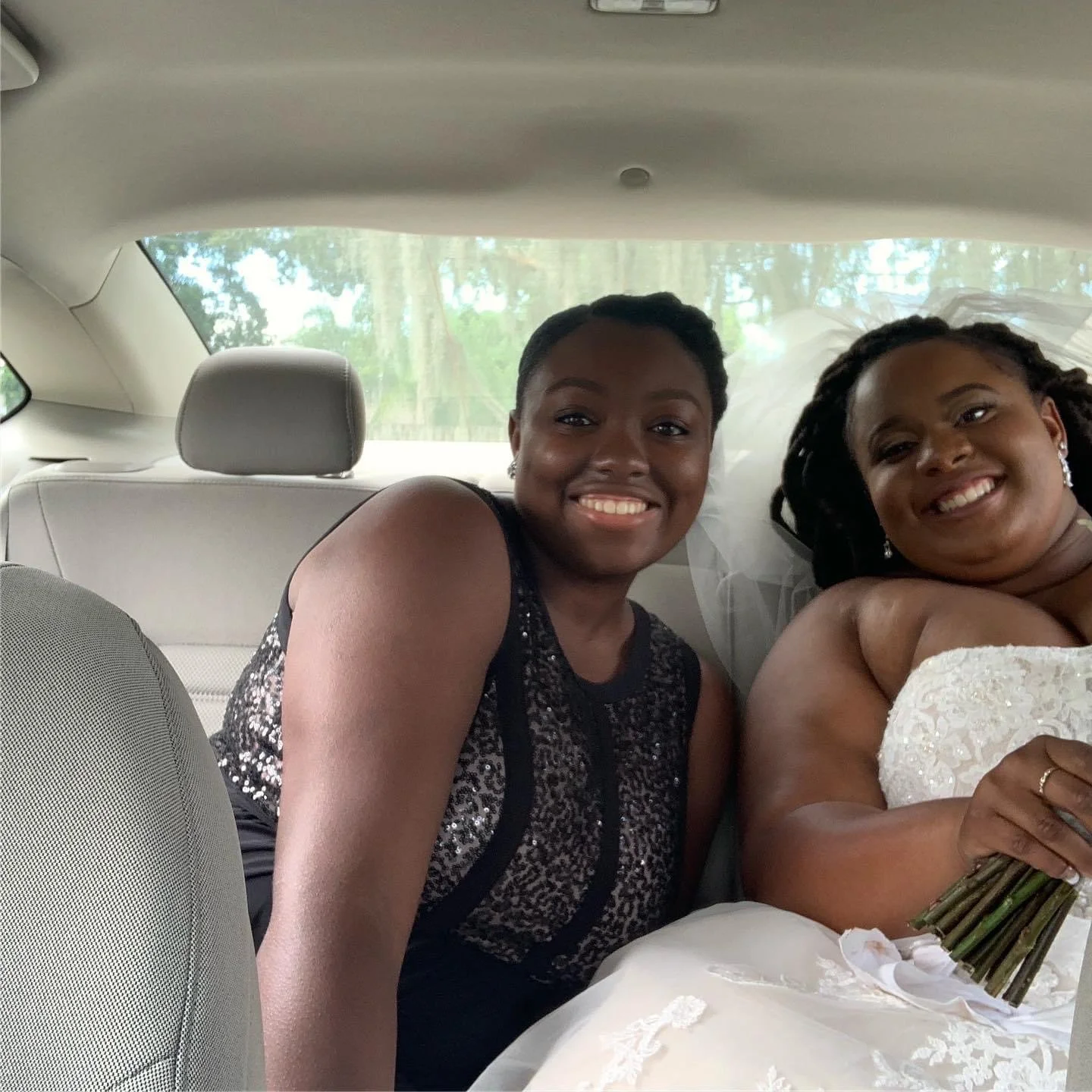 Two women sitting in the back seat of a car, smiling, with one wearing a black sleeveless dress with sequins and the other in a white wedding dress holding a bouquet of flowers, likely during a wedding.