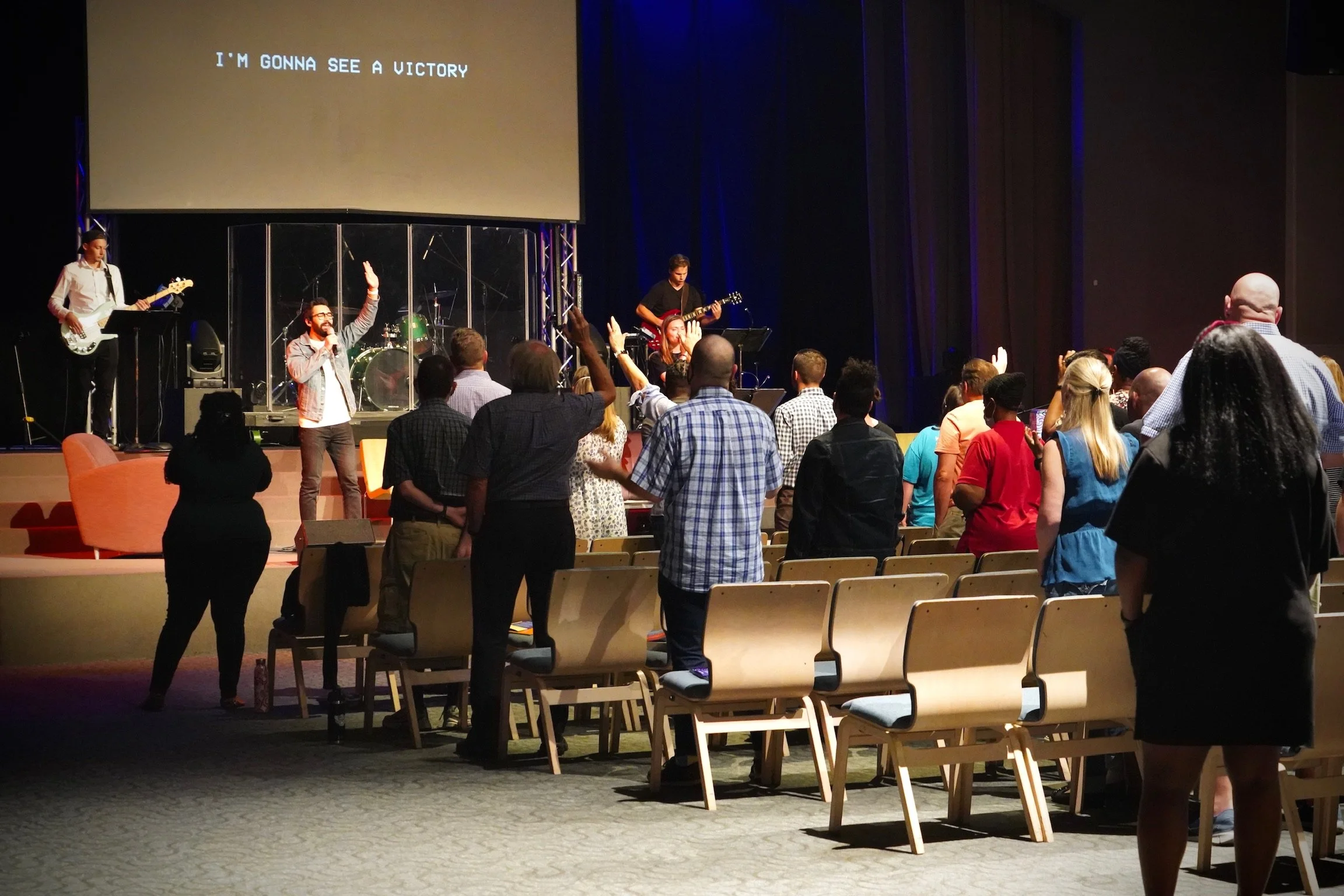 People gathered in a church or conference hall with a stage where a band is performing and a large screen displaying the words "I'M GONNA SEE A VICTORY." Some attendees are raising their hands while others are standing and facing the stage.