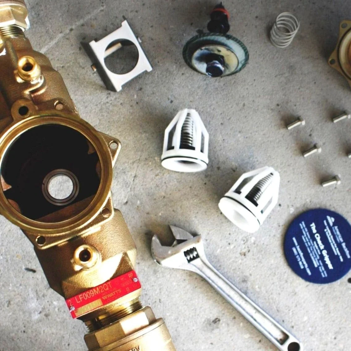 Plumbing components and tools on a concrete surface, including a brass valve, wrench, various fittings, springs, and small parts.