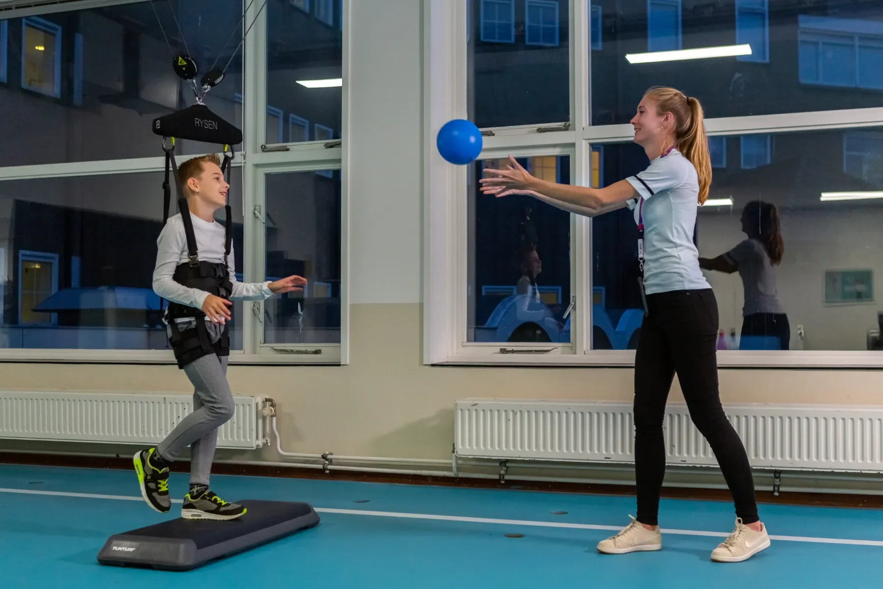 Niño realizando terapia de marcha con RYSEN y asistencia dinámica sobre suelo mientras la fisioterapeuta trabaja ejercicios funcionales con pelota. Bayber es distribuidor oficial en España y Portugal
