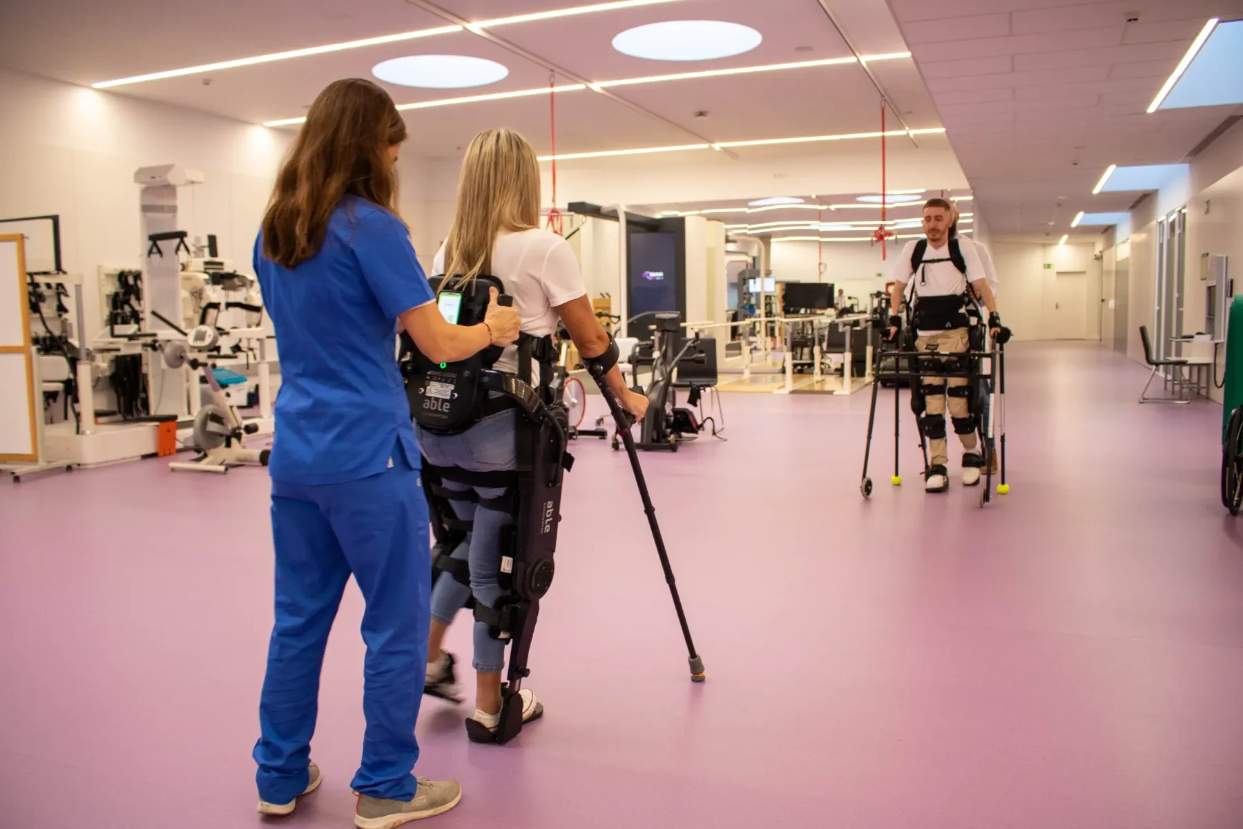 Dos pacientes utilizando ABLE Exoskeleton en sala de fisioterapia mientras el personal clínico supervisa la rehabilitación de la marcha. Contacta con Bayber para más información
