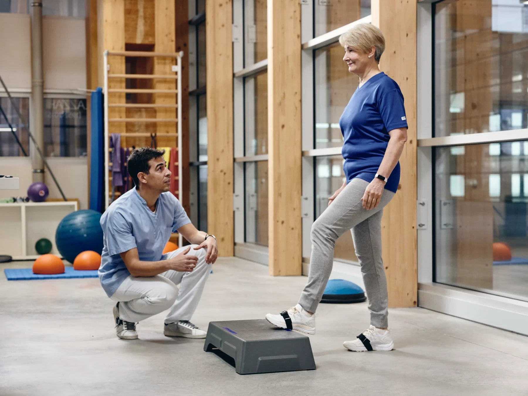 Paciente mayor entrenando el paso al escalón con sensores Euleria Lab en el tobillo mientras el fisioterapeuta supervisa la rehabilitación del equilibrio. Bayber es partner oficial en España y Portugal