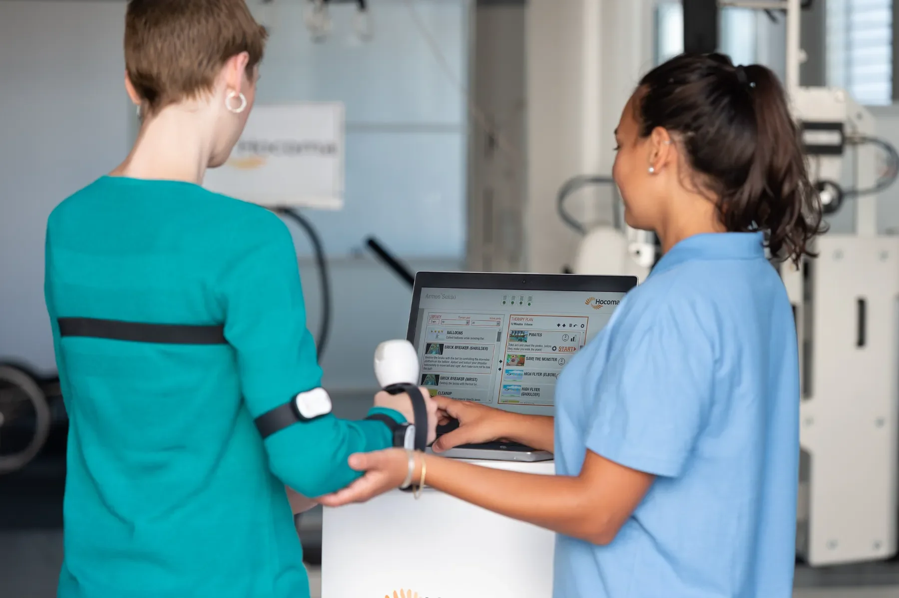 Paciente realizando rehabilitación de miembro superior con ArmeoSenso, usando sensores en brazo y pecho mientras la terapeuta ajusta el feedback aumentado en pantalla. Bayber es distribuidor oficial de Hocoma